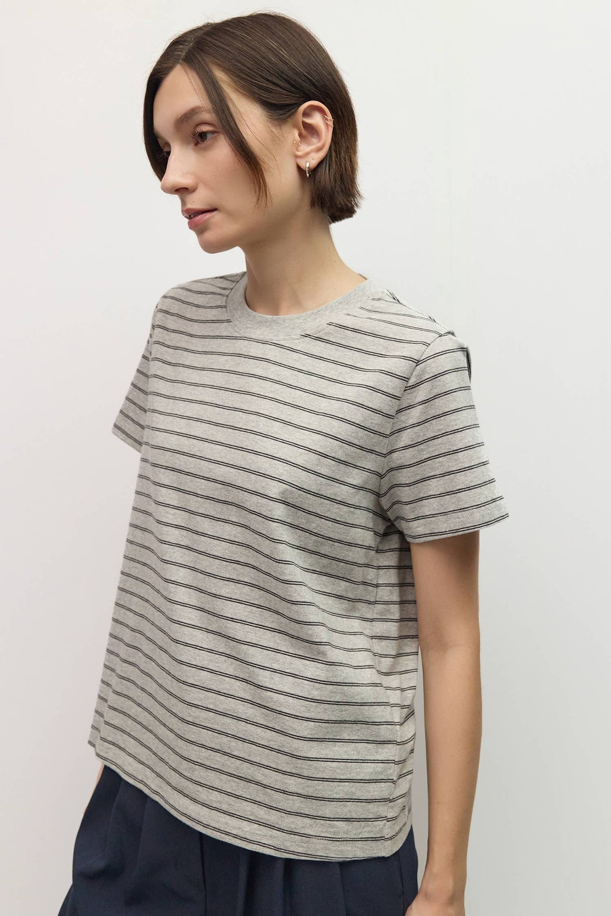 A woman wearing a short-sleeve crewneck t-shirt with thin horizontal gray stripes on a light gray background, paired with dark navy pants.