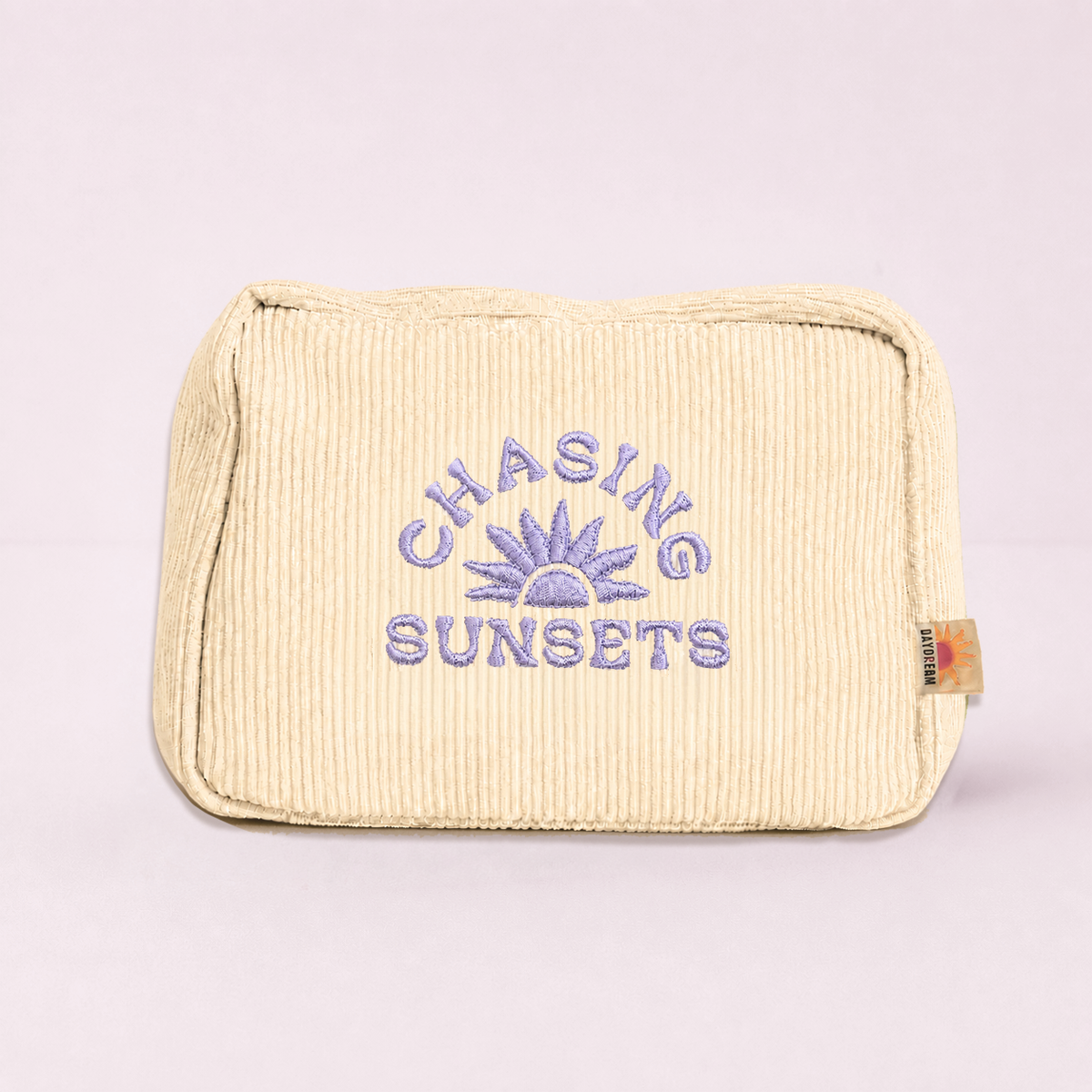 A cream-colored corduroy pouch with purple embroidered text reading 'CHASING SUNSETS' and a sun graphic, set against a solid light pink background.