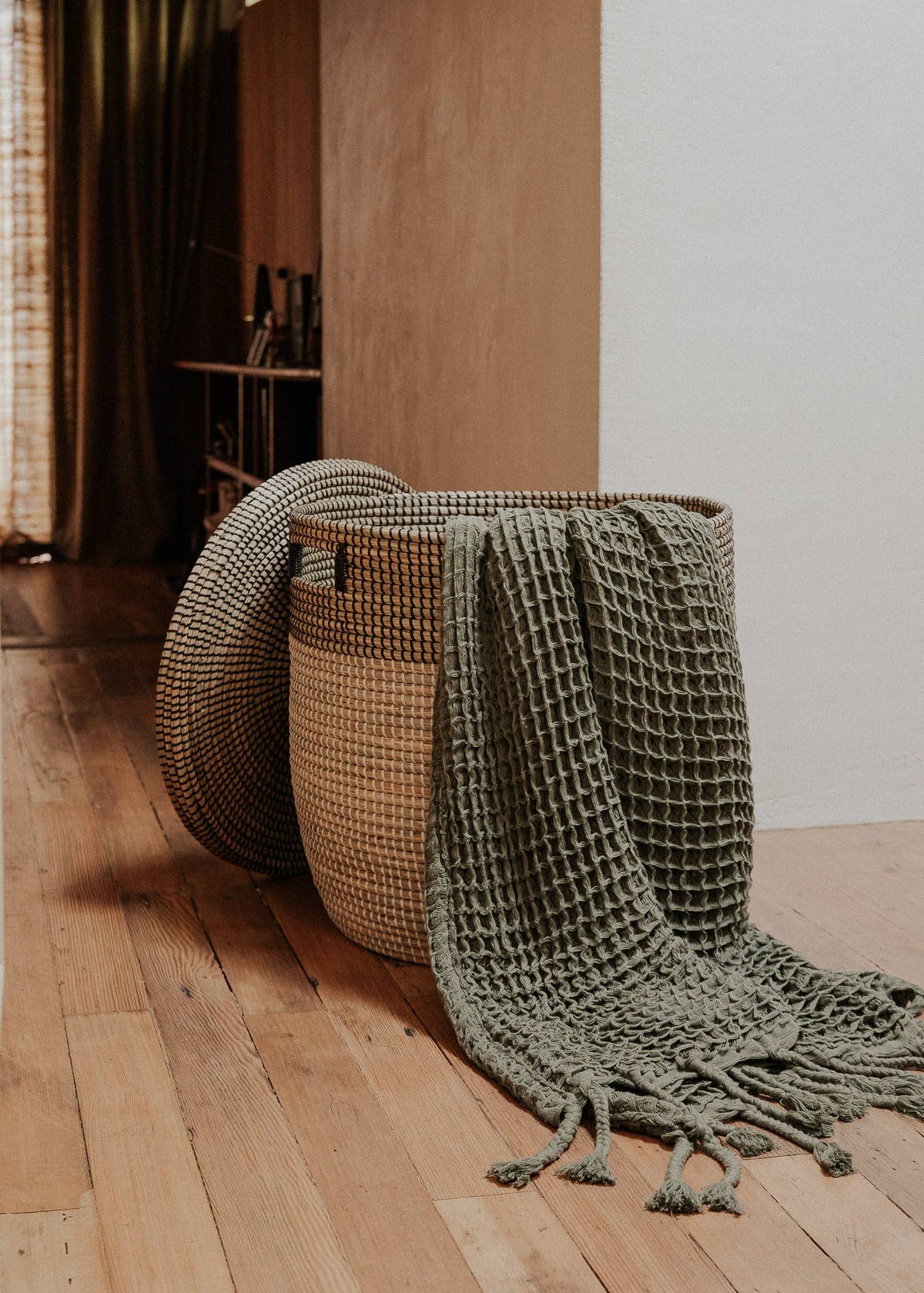 A woven round basket on a wooden floor, with a chunky knit throw blanket draped over it and a matching round woven mat leaning against the basket in a warm indoor setting.