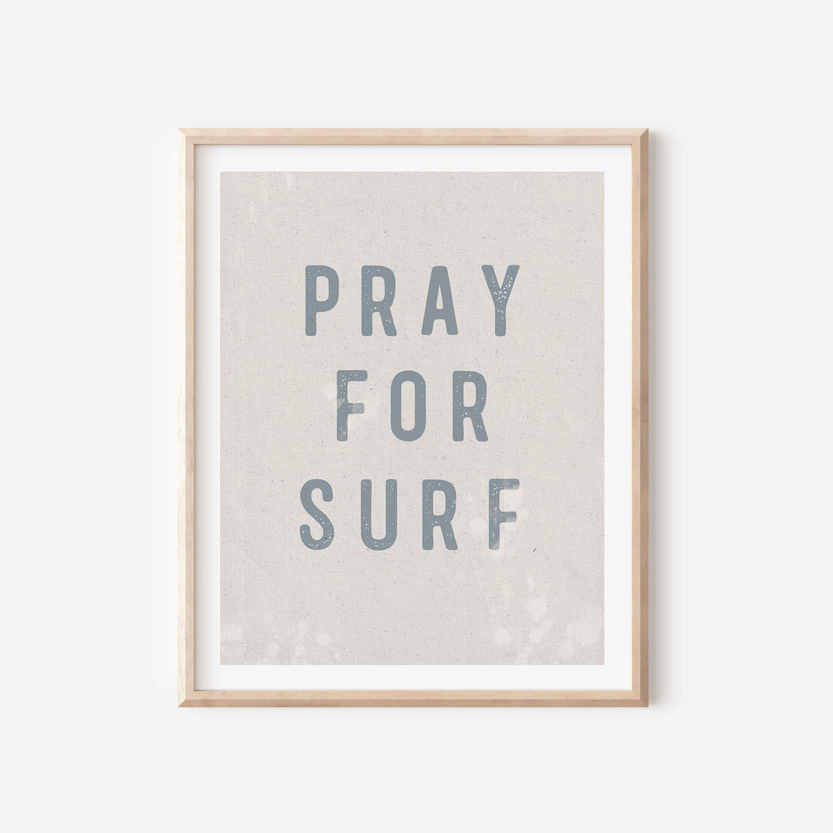Framed wall art print with the phrase 'PRAY FOR SURF' in muted blue-gray lettering on a light beige textured background, shown in a light wood frame against a white wall.