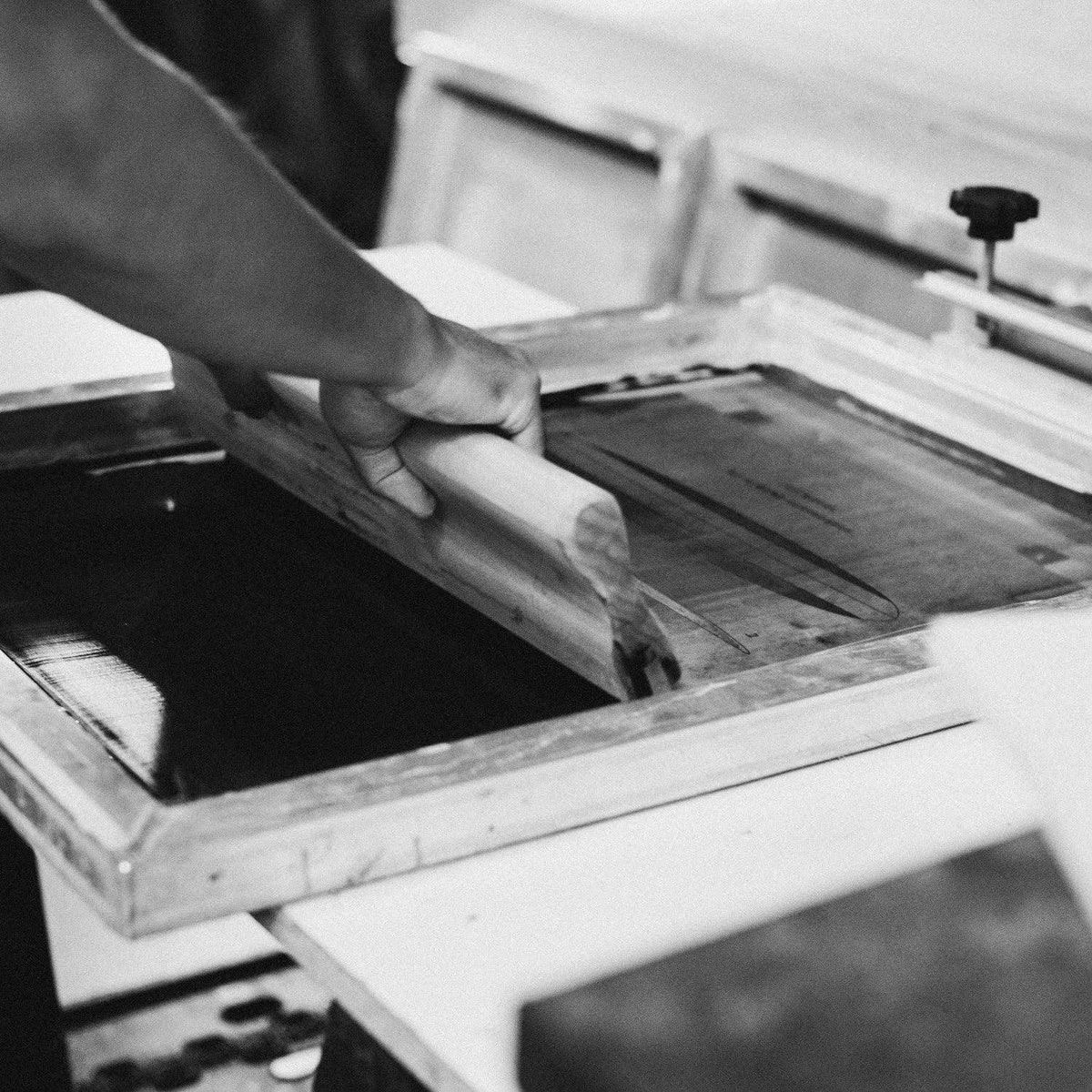 A black and white photograph showing a person's arm and hand pushing a wooden screen squeegee over a printing frame containing ink.