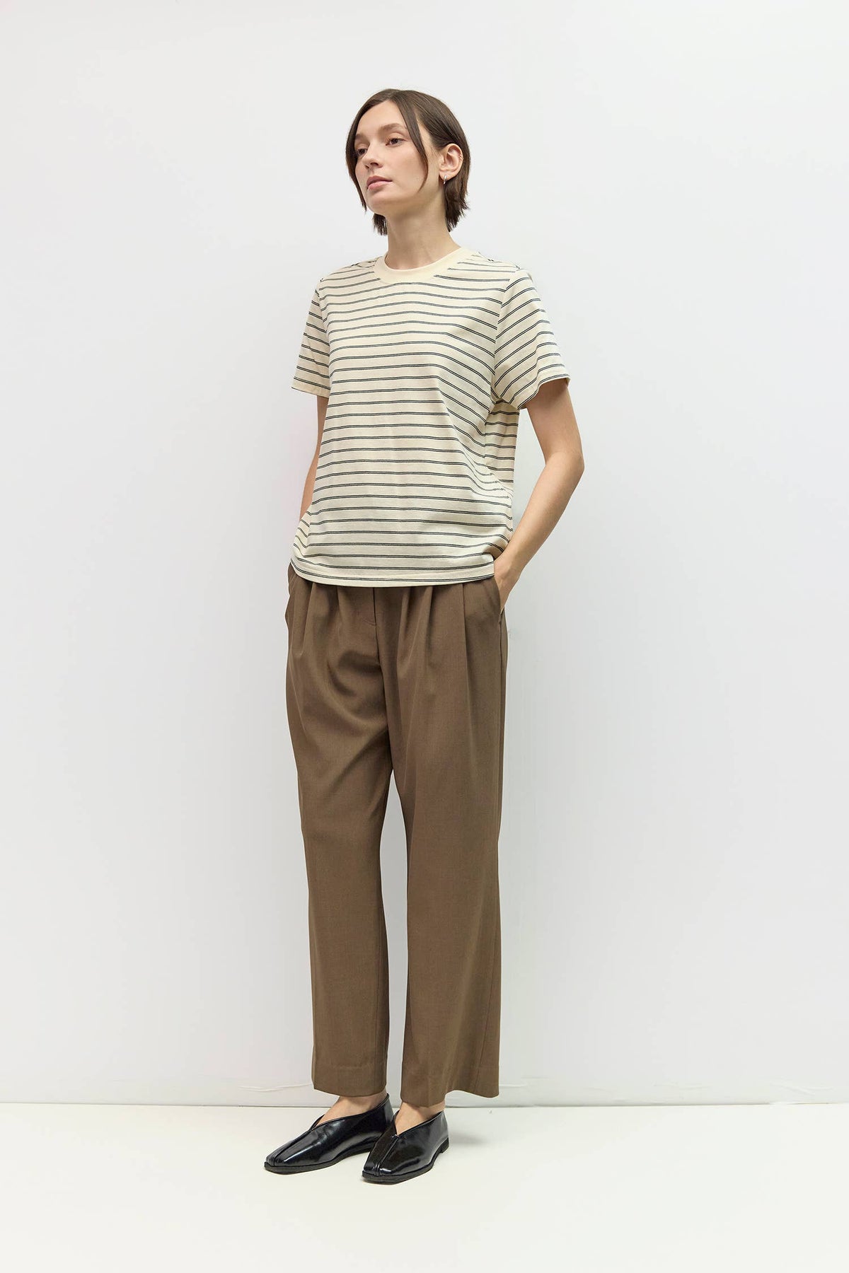 A woman modeling a short-sleeve crewneck t-shirt with thin beige and brown horizontal stripes, paired with brown wide-leg trousers and black leather loafers, standing against a plain white studio background.