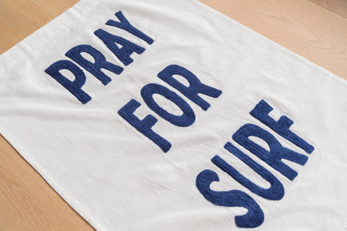 A white towel laid flat on a light wood surface, featuring the phrase 'PRAY FOR SURF' in large navy blue letters.