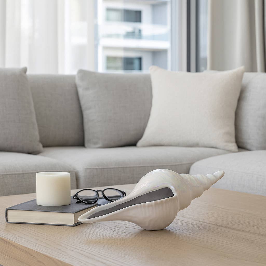 A white ceramic conch shell decorative object resting on a light wood table next to a book, a candle, and eyeglasses in a modern living room setting.