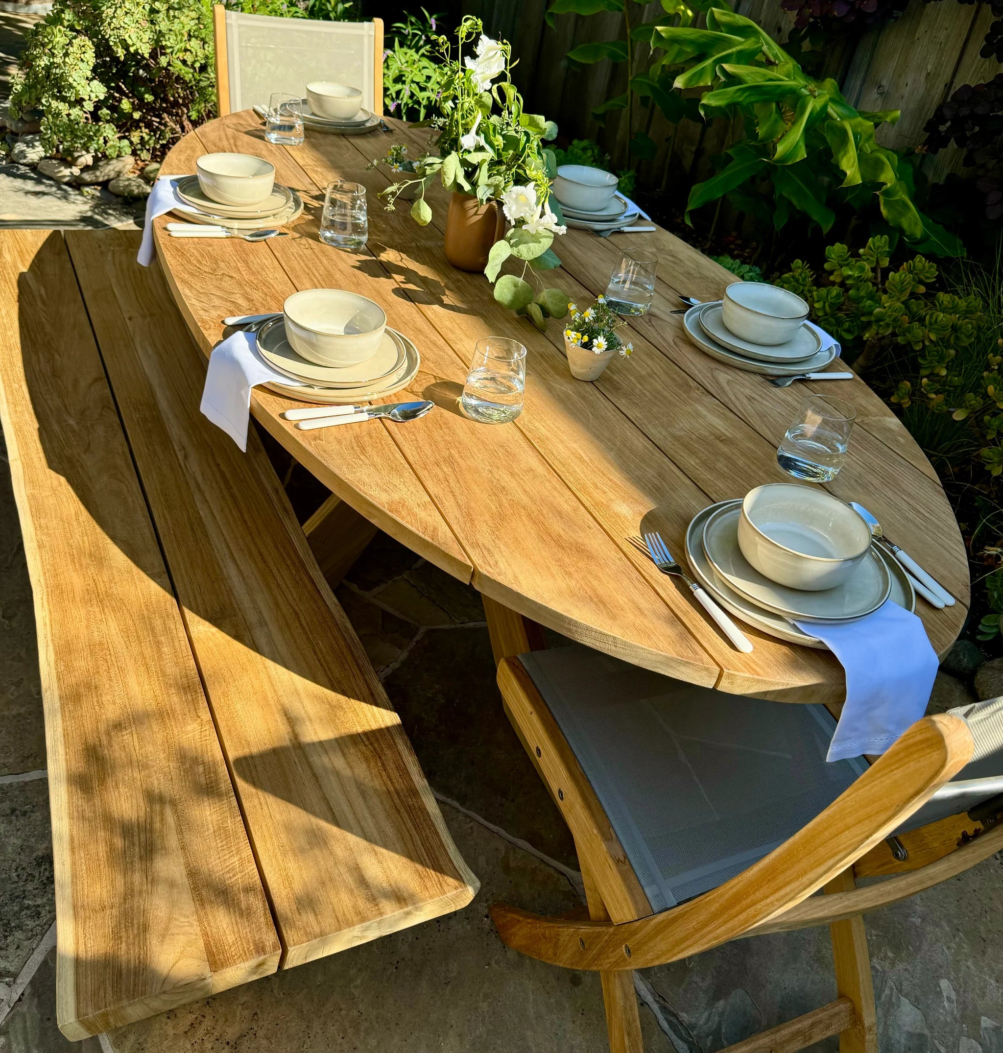 Wooden outdoor dining table set with plates, glasses, and cutlery in a garden setting.