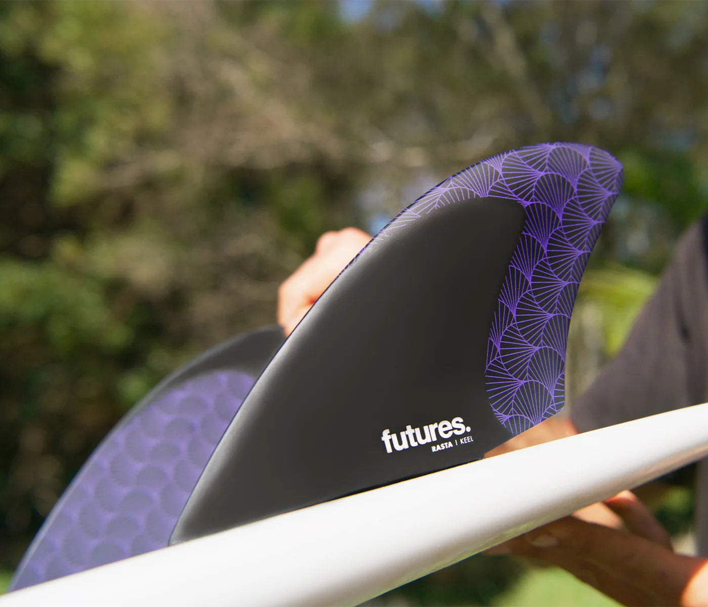 Person holding a surfboard with purple Futures Dave Rastovich Rasta Honeycomb Keel Fin Set, against a blurred natural background.