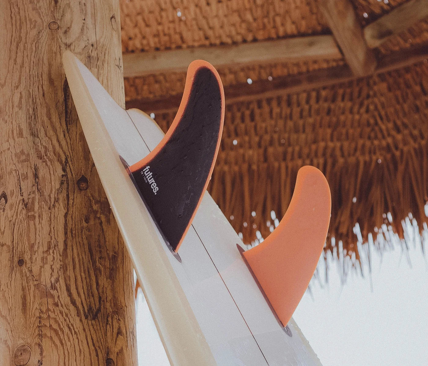 Close-up of a surfboard with tFutures T2 Fiberglass Twin Fin Set attached, leaning against a wooden post under a thatched roof. One fin is dark charcoal/black with orange trim and visible 'Rudder' branding, and the other is solid orange.