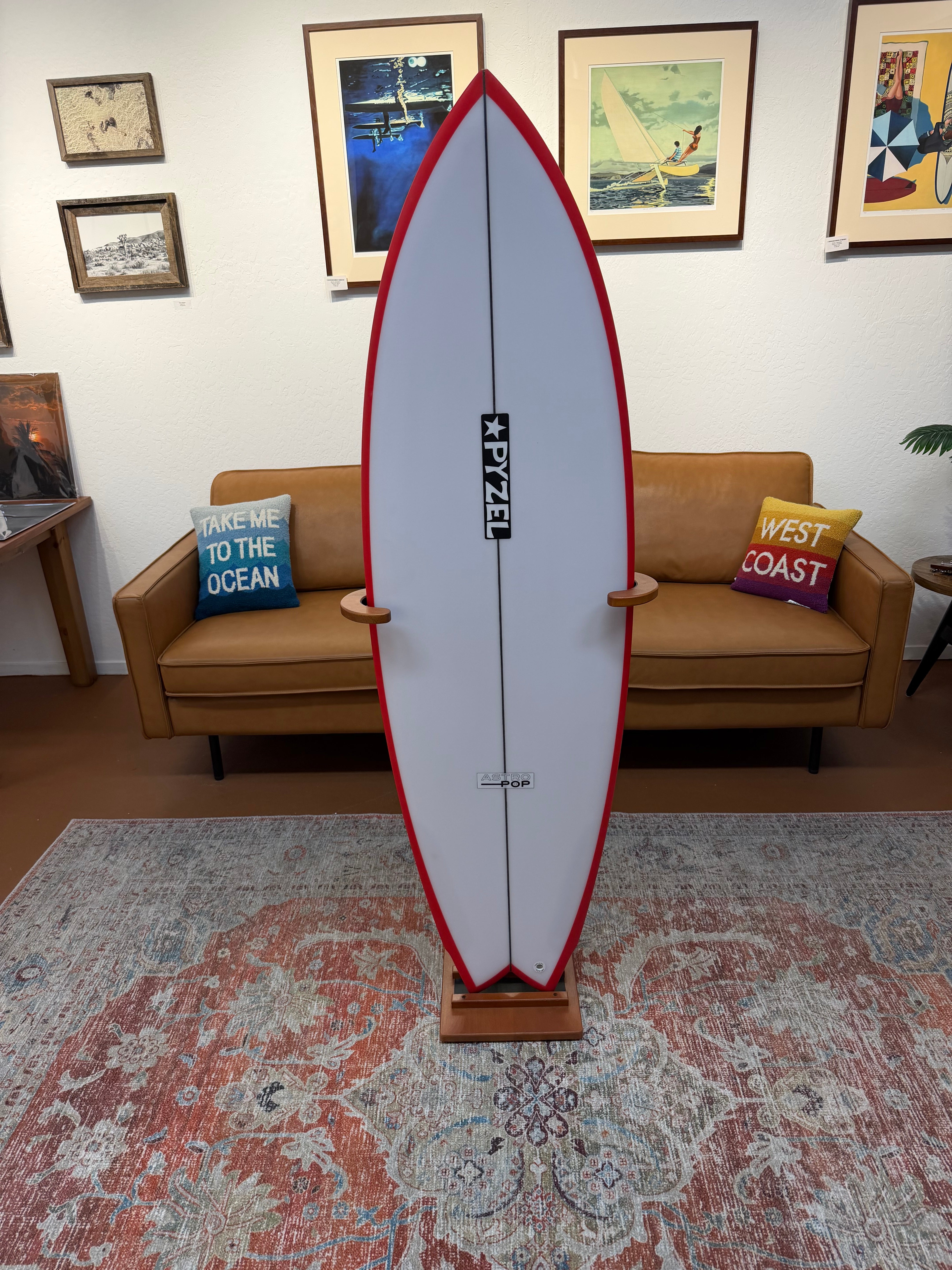 A freestanding surfboard-shaped decor piece or display board with a white center, red border, and black vertical logo, shown on a wooden stand in a living room setting with a tan sofa, framed wall art, and a patterned rug.