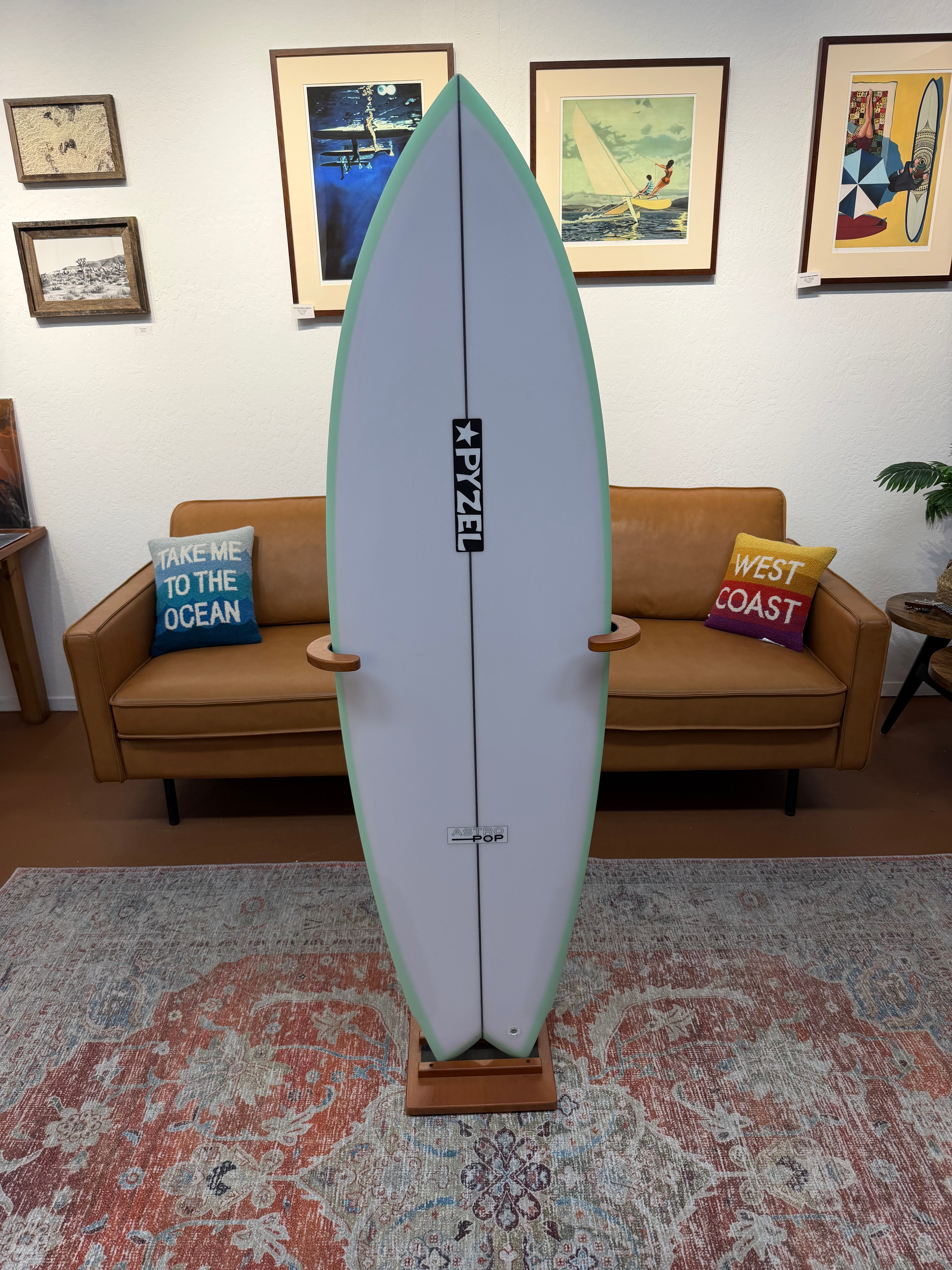 A freestanding surfboard on a wooden display stand in a living room setting, with a tan sofa, framed wall art, and a patterned rug behind it. The surfboard is light gray with a mint green edge and a black central logo reading 'FYEZ'.