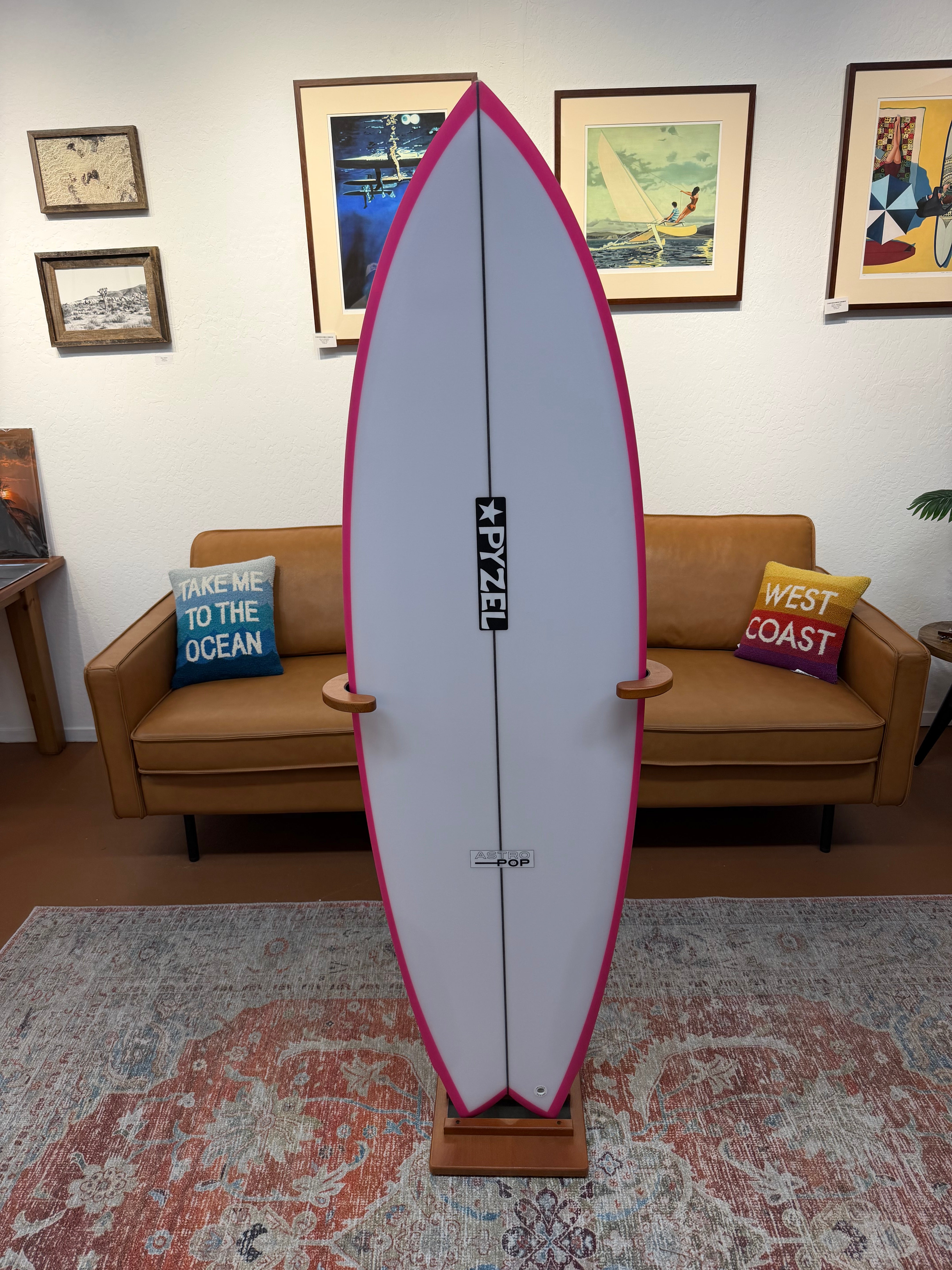 A Pyzel surfboard displayed vertically on a stand in a living room setting with a brown sofa, framed wall art, and a patterned rug.