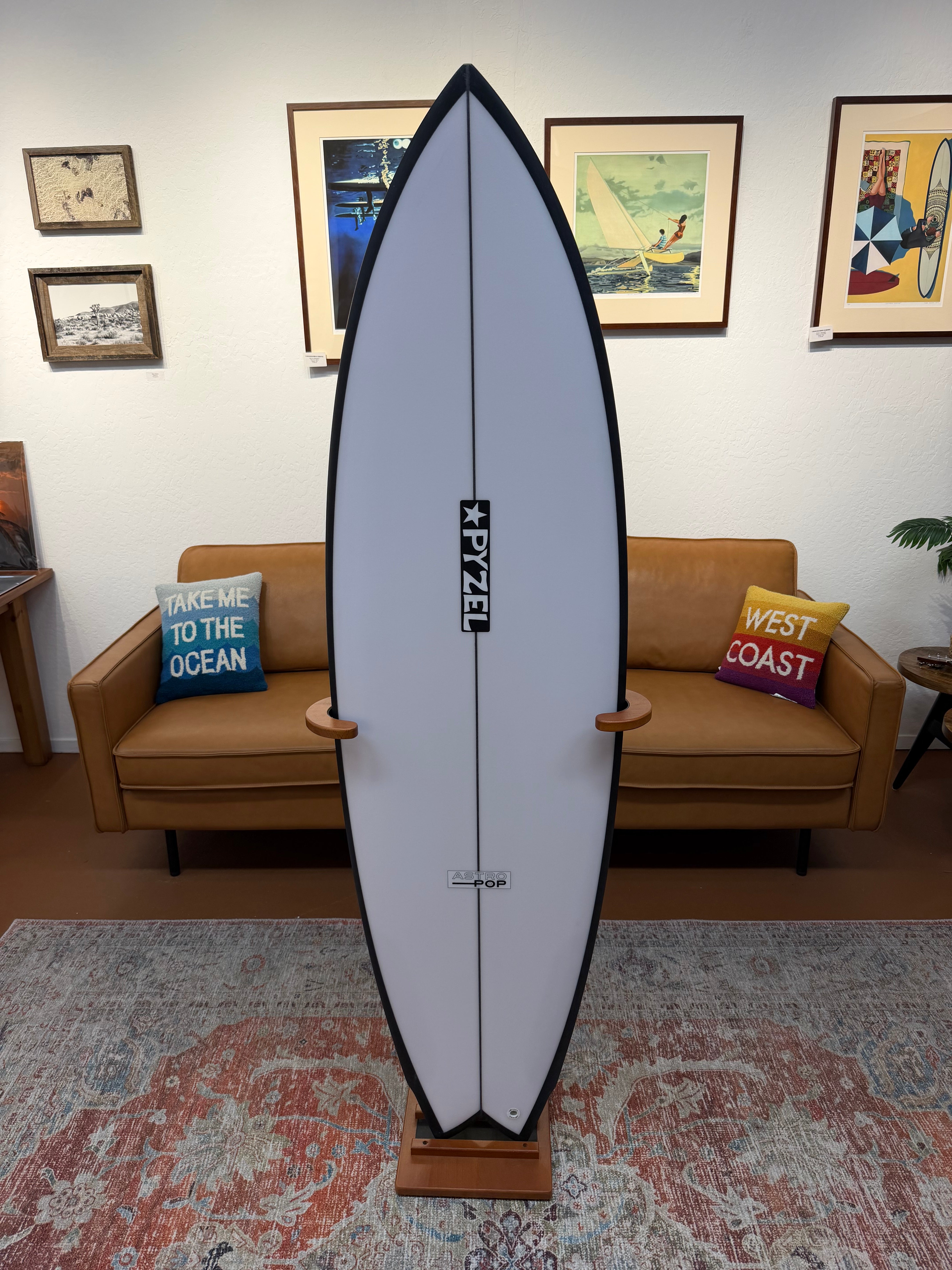A white surfboard with a black outline and a vertical black 'KAYZE' logo, displayed on a wooden stand in a living room setting with a tan leather sofa and framed wall art in the background.