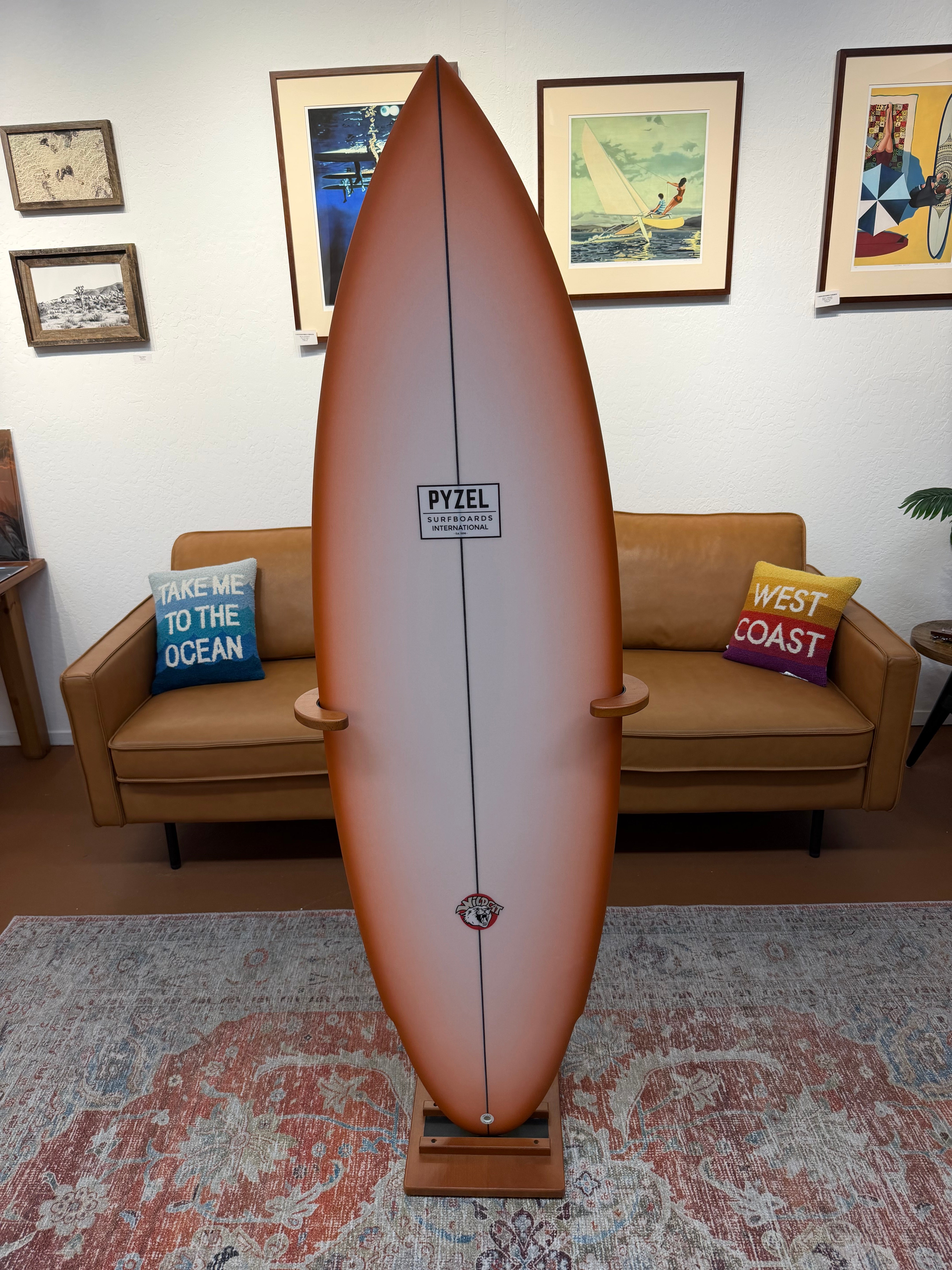 A vintage-style PYZEL surfboard displayed upright on a wooden stand in a living room setting, with a tan leather sofa and framed wall art in the background.