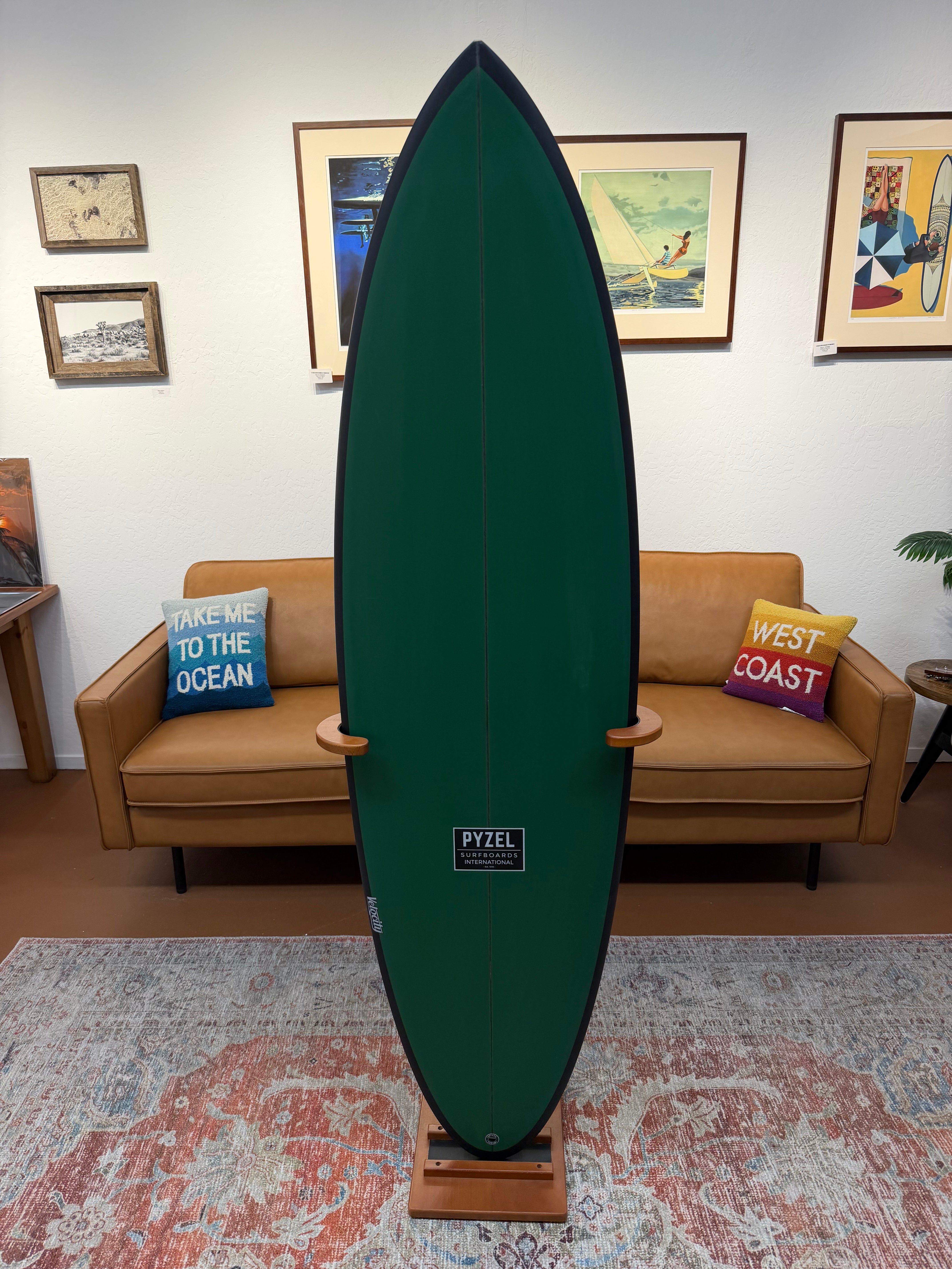 A dark green Pyzel surfboard displayed upright on a wooden stand in a living room setting with a tan sofa, framed wall art, and a patterned rug.