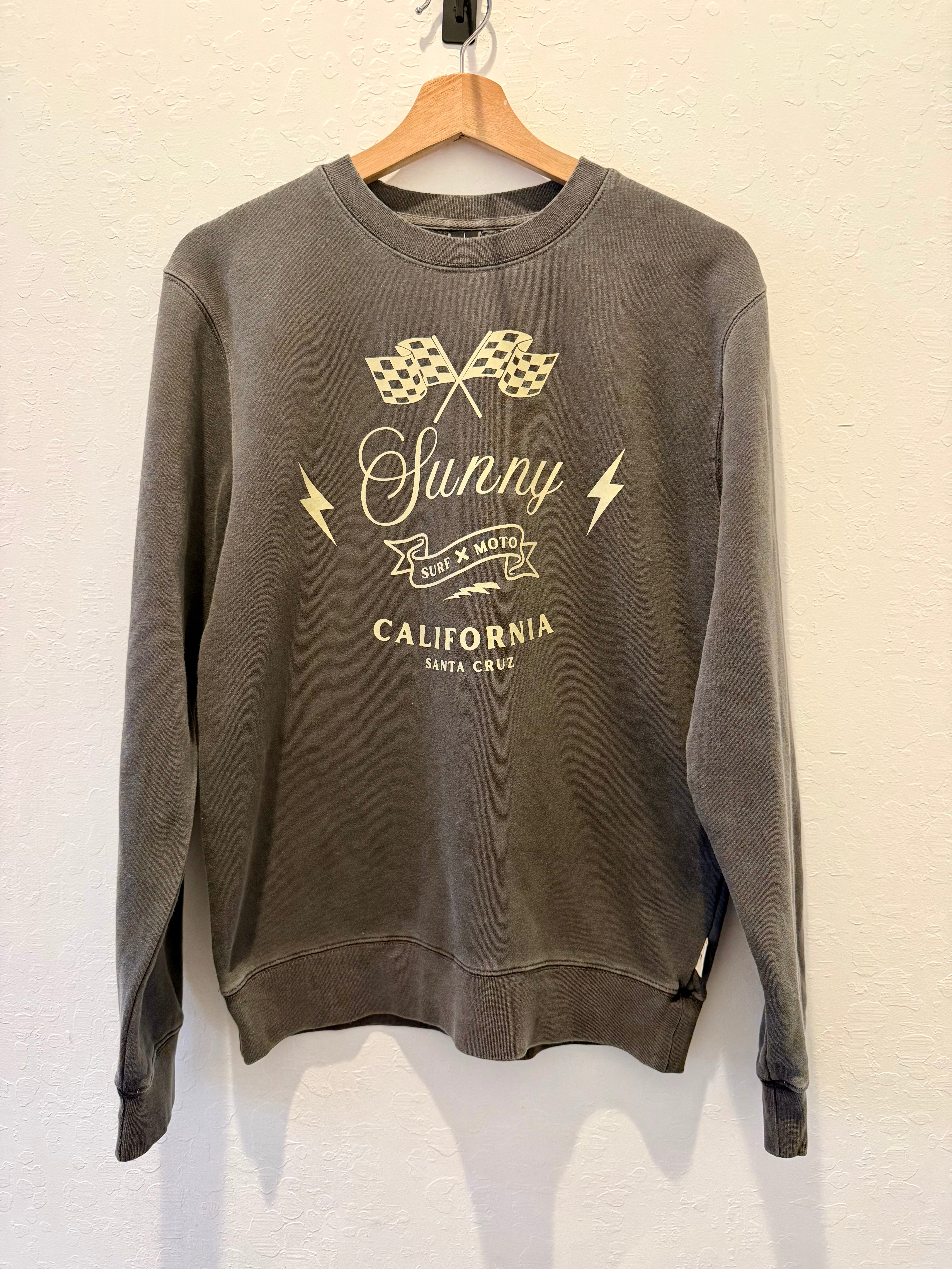 A dark gray washed crewneck sweatshirt hanging on a wooden hanger against a textured off-white wall. The sweatshirt features a vintage-style graphic print in a light beige color with checkered flags, lightning bolts, and the text 'Sunny Car & Auto' and 'CALIFORNIA SANTA CRUZ'.