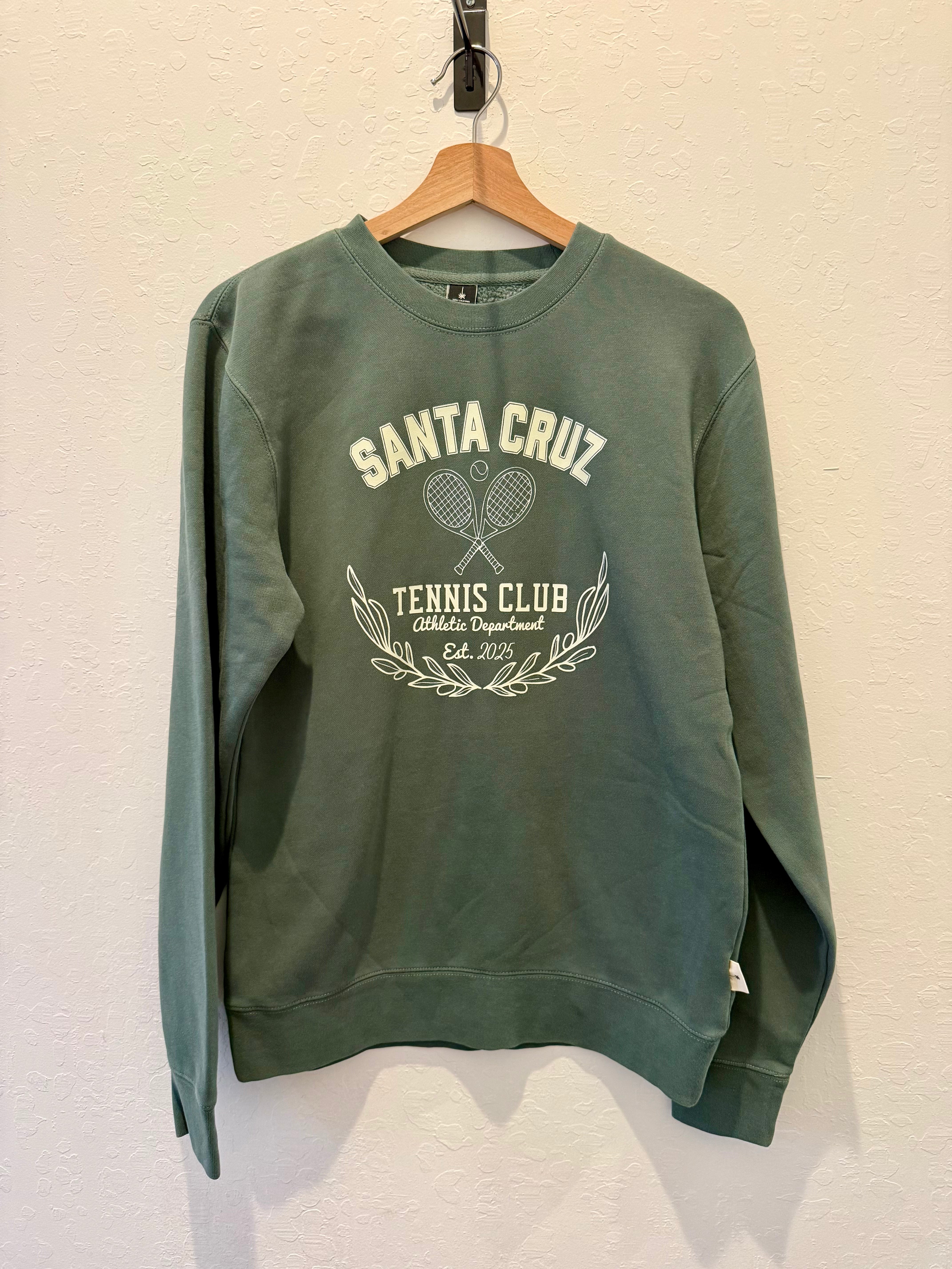 A sage green crewneck sweatshirt hanging on a wooden hanger against a textured off-white wall. The sweatshirt features a cream-colored graphic on the front that reads 'SANTA CRUZ TENNIS CLUB' with crossed tennis rackets and laurel leaves.