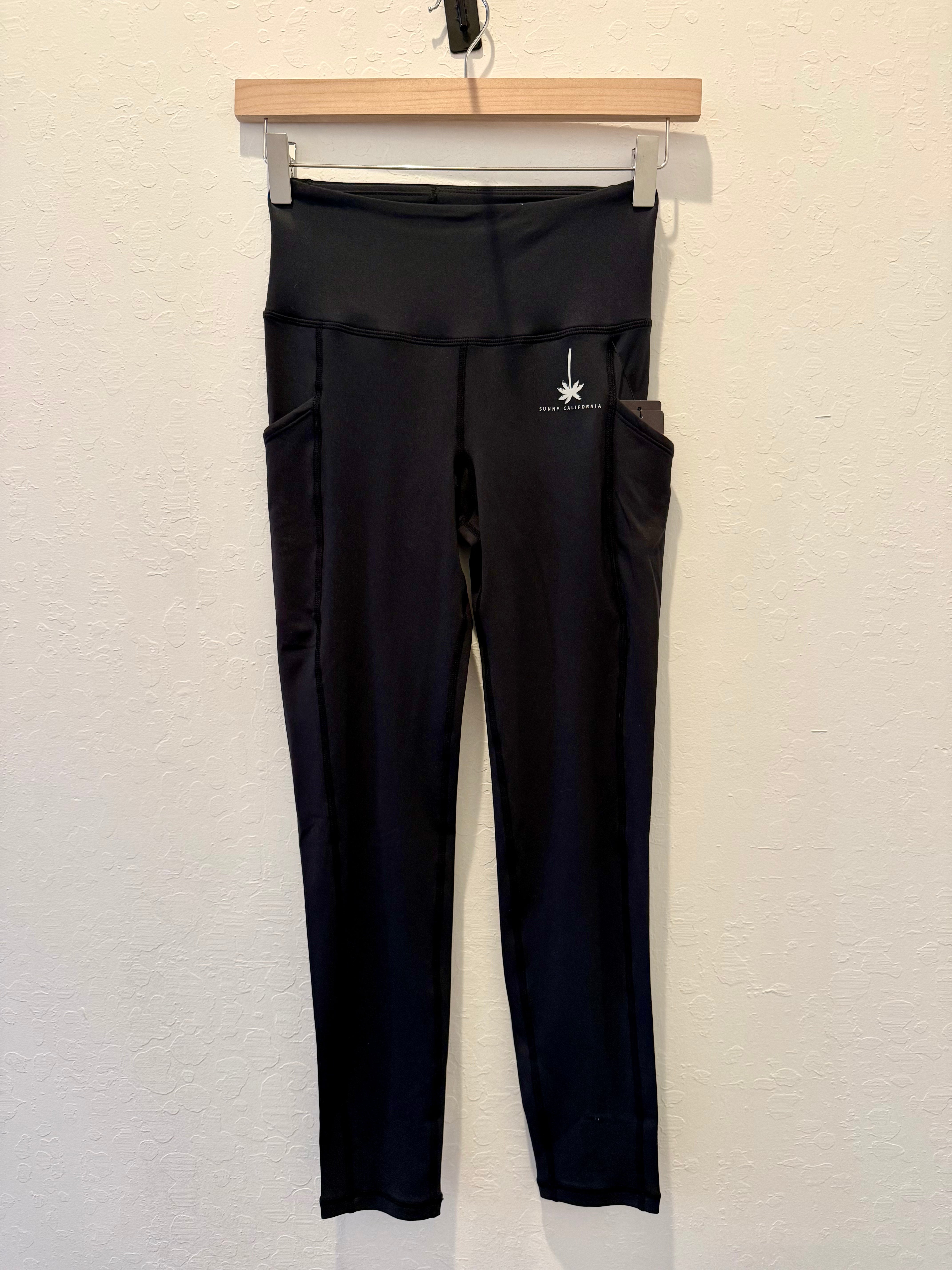 Black high-waisted activewear leggings with side pockets, hanging on a wooden hanger against a textured off-white wall. A small white logo is visible on the upper right hip.