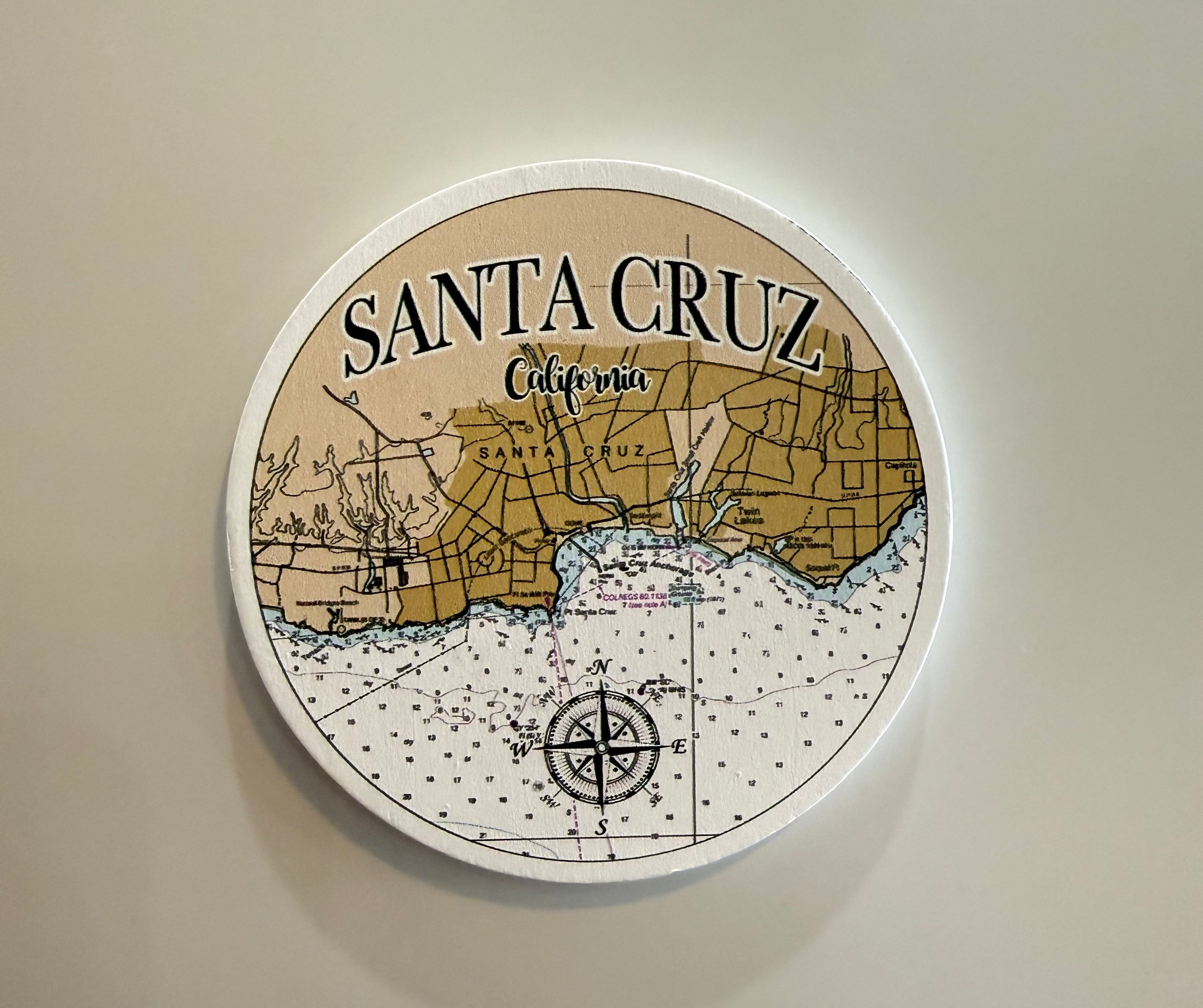 A round white-framed wall art print featuring a vintage-style map of Santa Cruz, California, with black text and linework on a beige and tan background, mounted on a plain light wall.