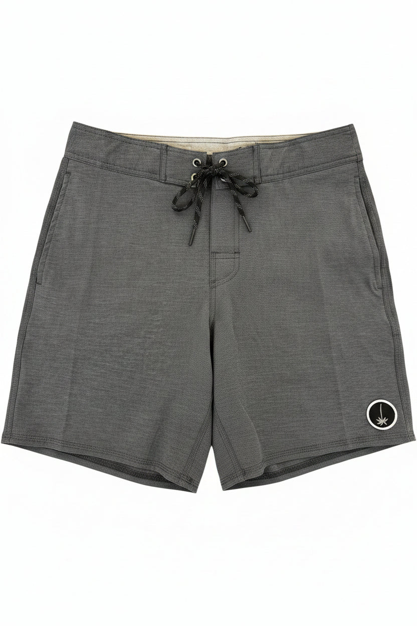 Dark gray heathered board shorts with a black drawstring and a small white circular logo patch on the lower left leg, laid flat on a wooden floor with yellow retail tags attached.