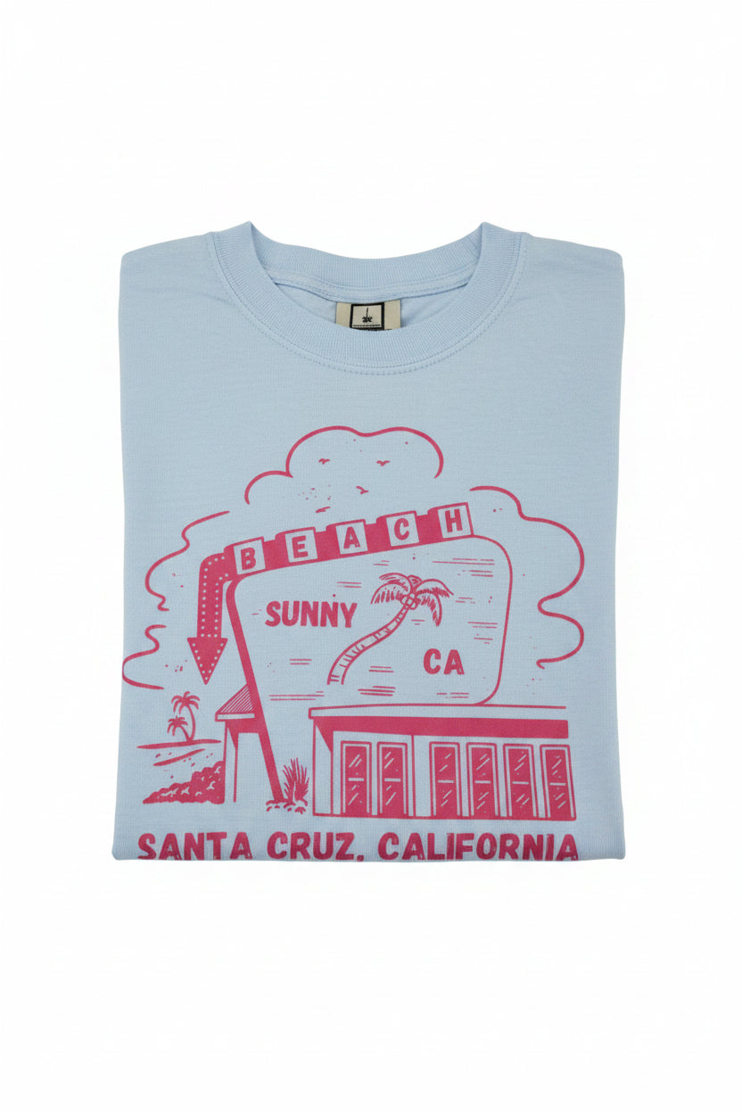 A folded light blue t-shirt with a red graphic print of the 'Sunny Beach' sign in Santa Cruz, California, displayed on a reflective surface.