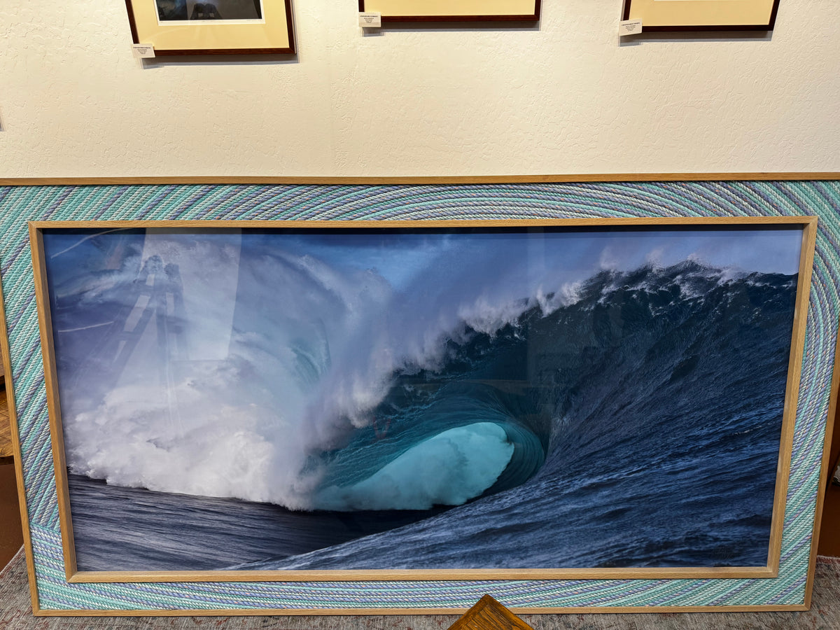 A large framed ocean wave photograph leaning against a wall in an indoor gallery or showroom. The print shows a dramatic blue and white curling wave, surrounded by a patterned blue mat and a light wood frame.