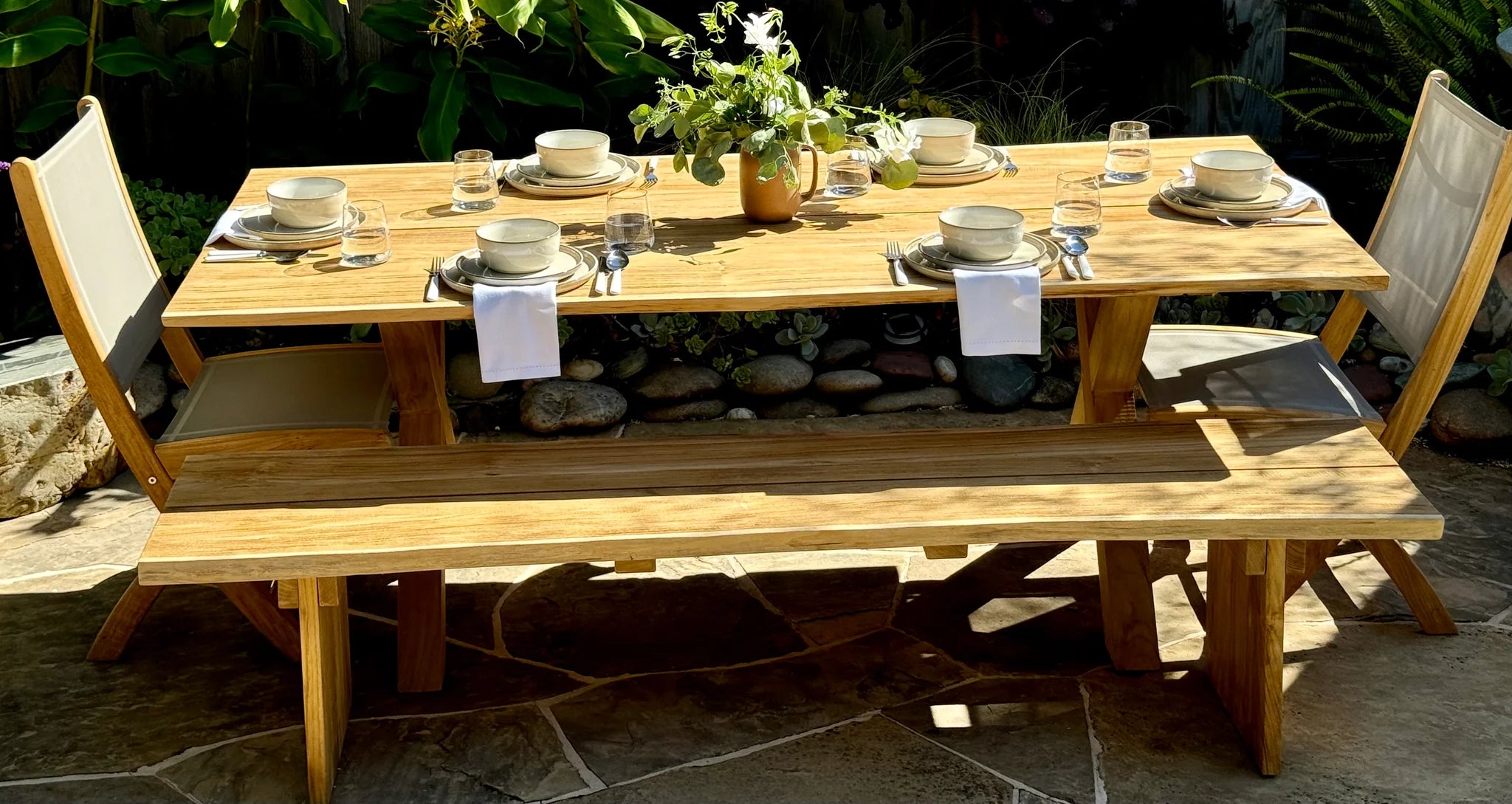 Wooden outdoor dining table set with chairs and tableware on a stone patio.