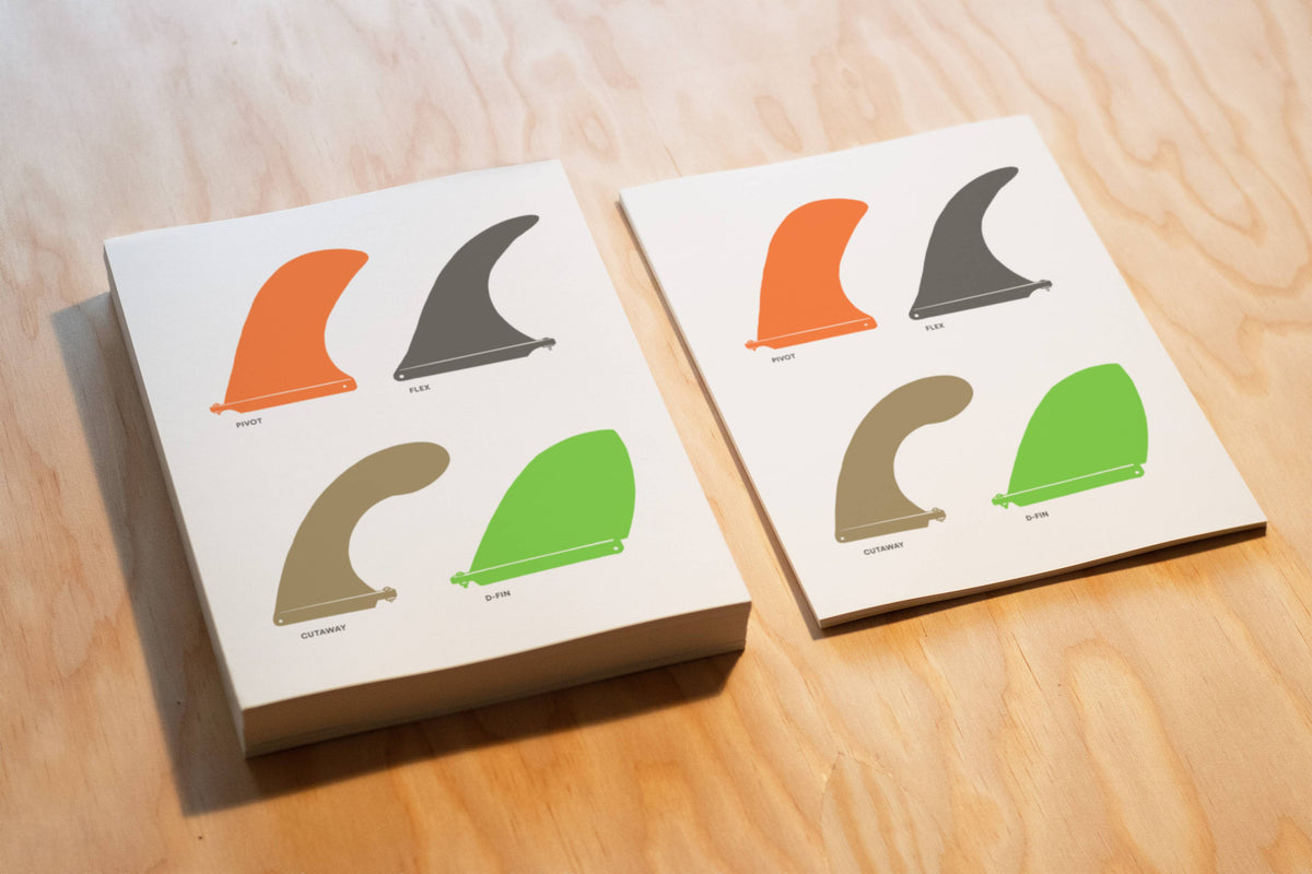 Two square printed cards or boxes on a light wood surface, each showing four surfboard tail fin illustrations in orange, dark gray, tan, and green with small labels underneath.