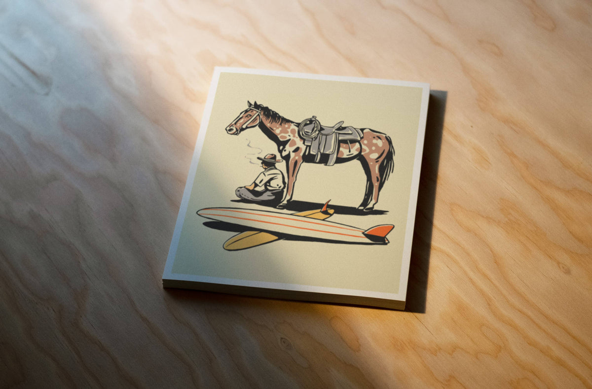 A square art print featuring an illustration of a horse with a rider and a surfboard, displayed on a light wood grain surface under warm directional sunlight.