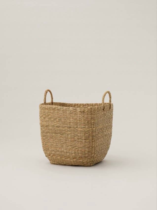 A natural woven straw storage basket with two loop handles, shown on a neutral light gray background.