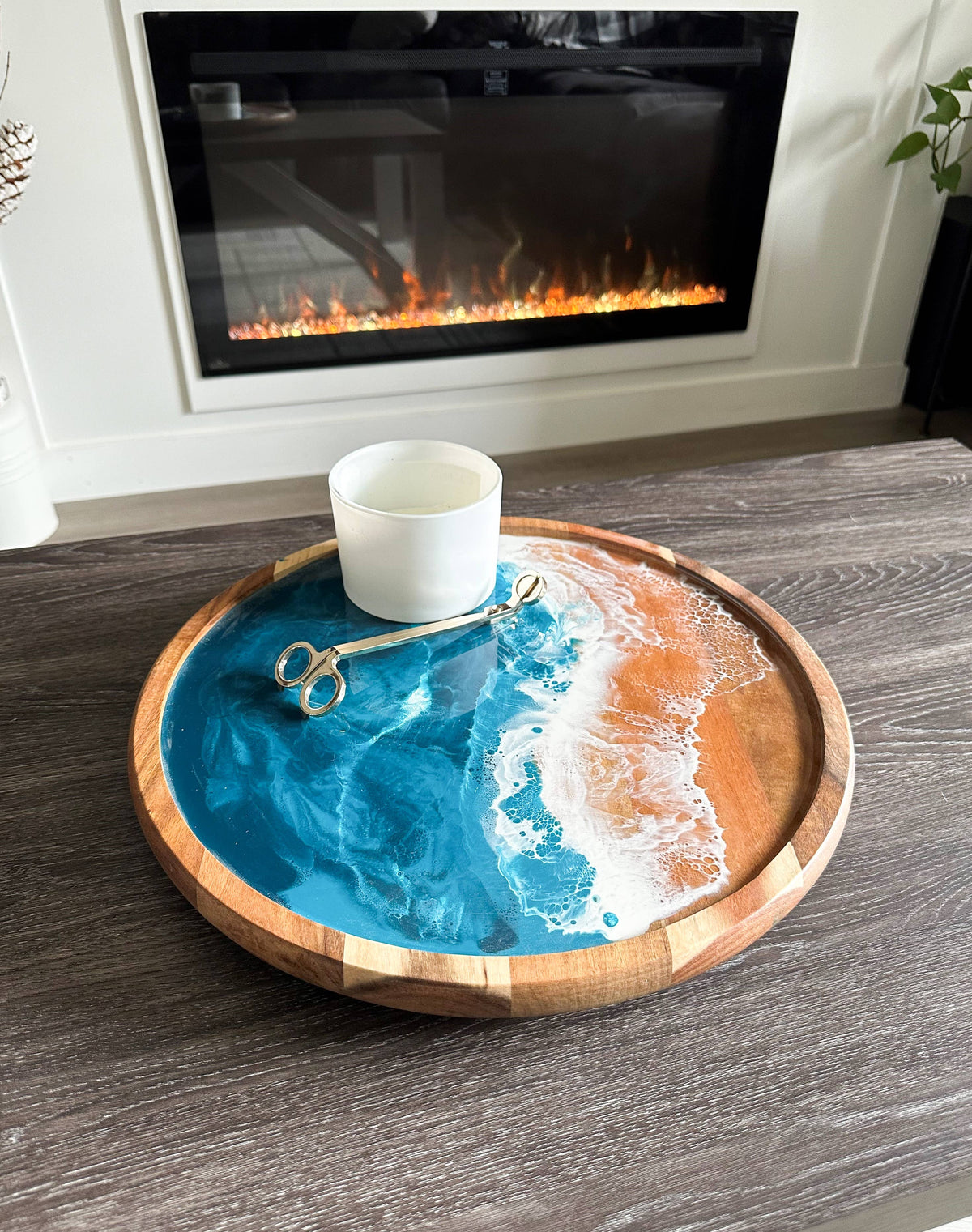 A round wooden tray with a resin ocean design in shades of blue, white, and orange, styled on a dark wood table with a white candle and gold scissors in front of a fireplace.
