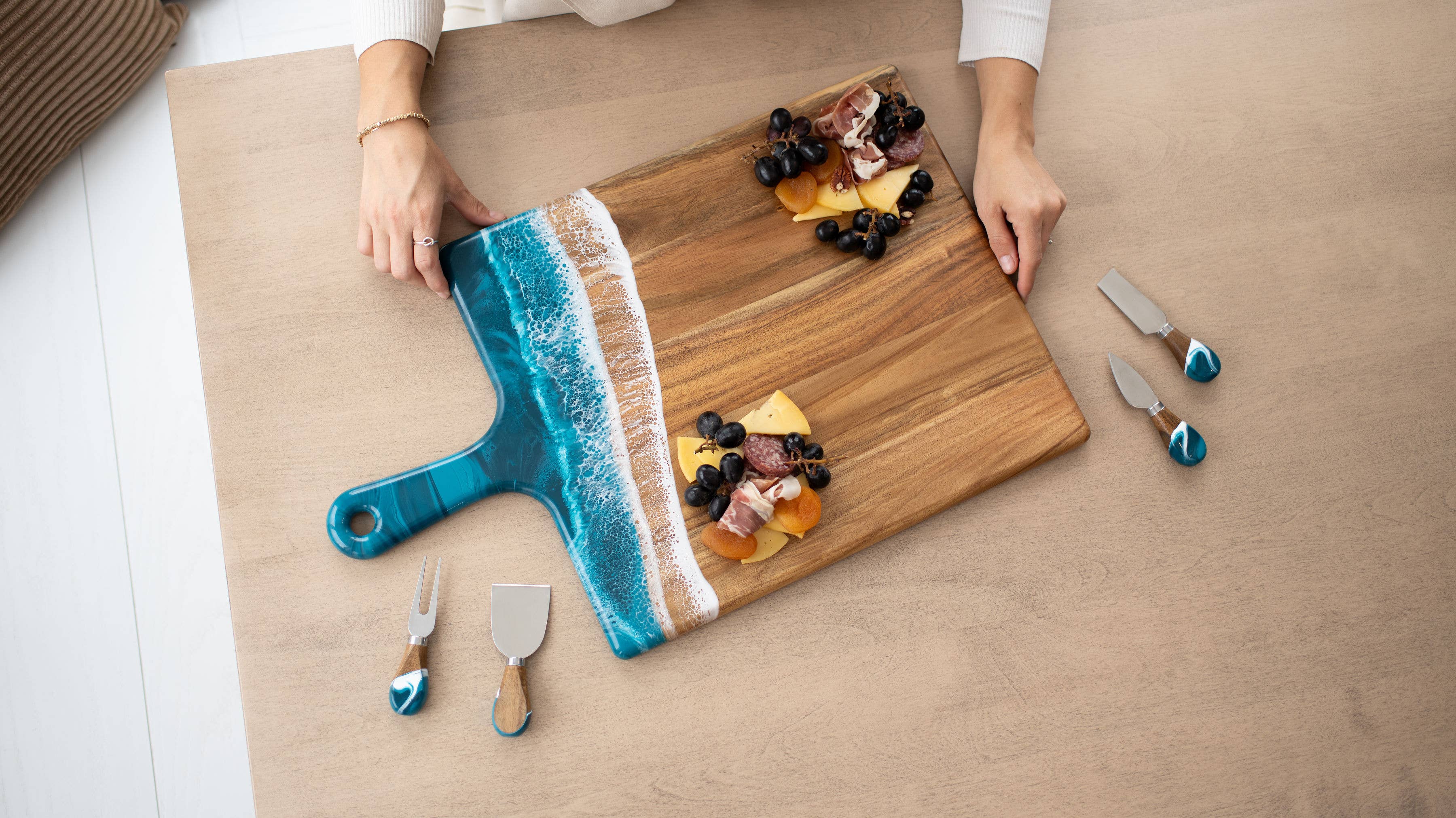Person holding a decorative cutting board with a blue handle, surrounded by small knives and food items on a wooden surface.