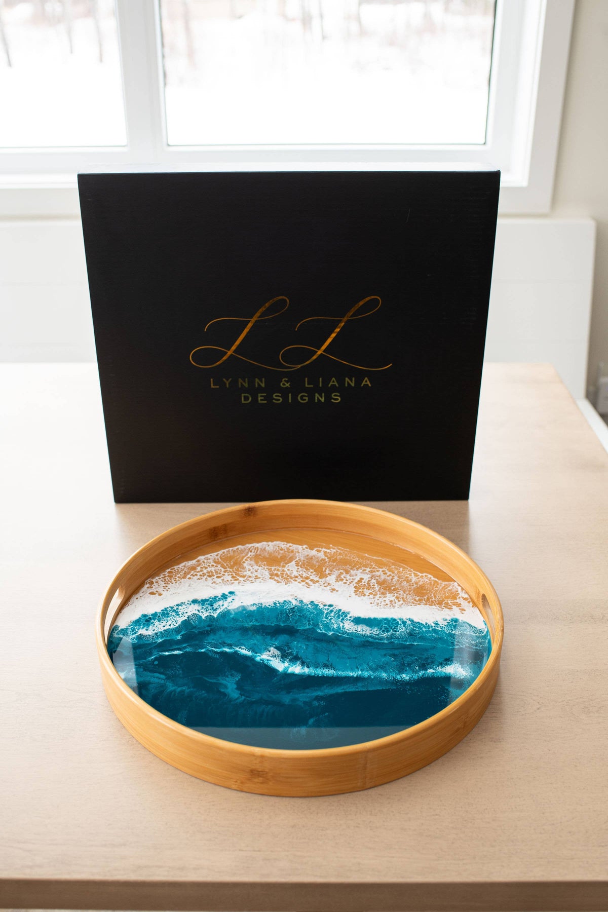 A round wooden tray filled with a resin ocean wave design in shades of blue and white, displayed on a light wood table in front of a black branded box that reads 'Lynn & Liana Designs'.
