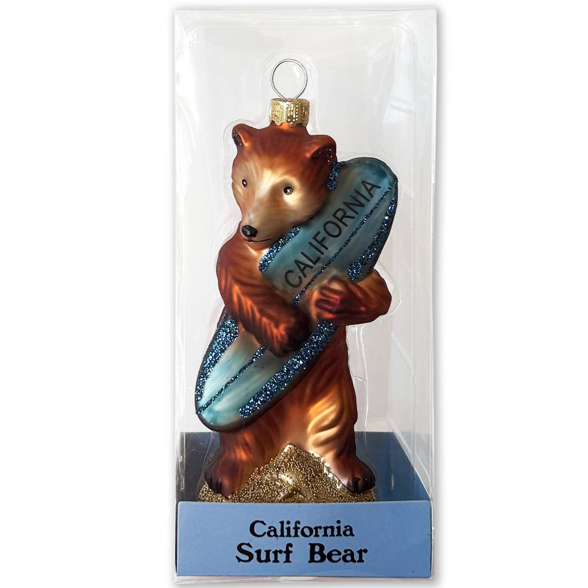 A Christmas ornament shaped like a brown bear surfing on a blue surfboard with 'CALIFORNIA' printed on it, packaged in a clear plastic sleeve with a blue base labeled 'California Surf Bear'.