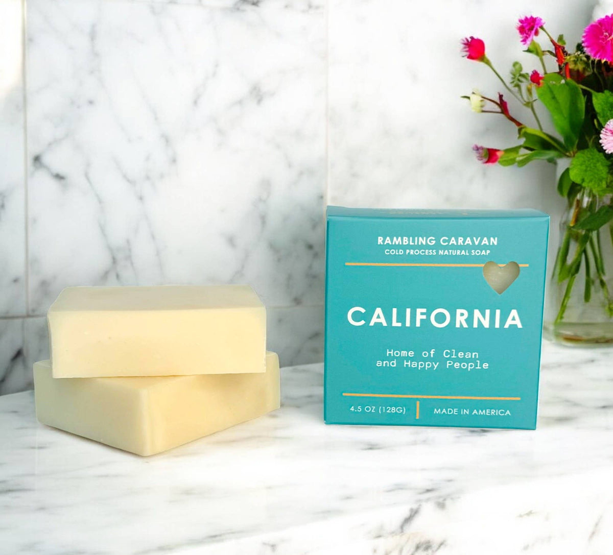 A bar of natural soap and its turquoise box on a white marble surface, with a vase of pink flowers in the background. The box reads 'RAMSLING CARAVAN' and 'CALIFORNIA'.