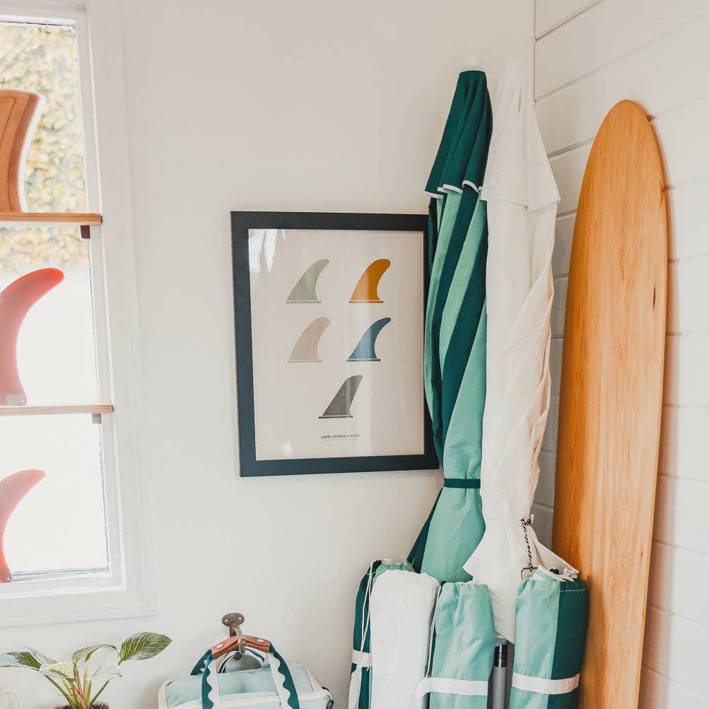 Surfboard in a case, umbrella, and framed artwork on a wall.