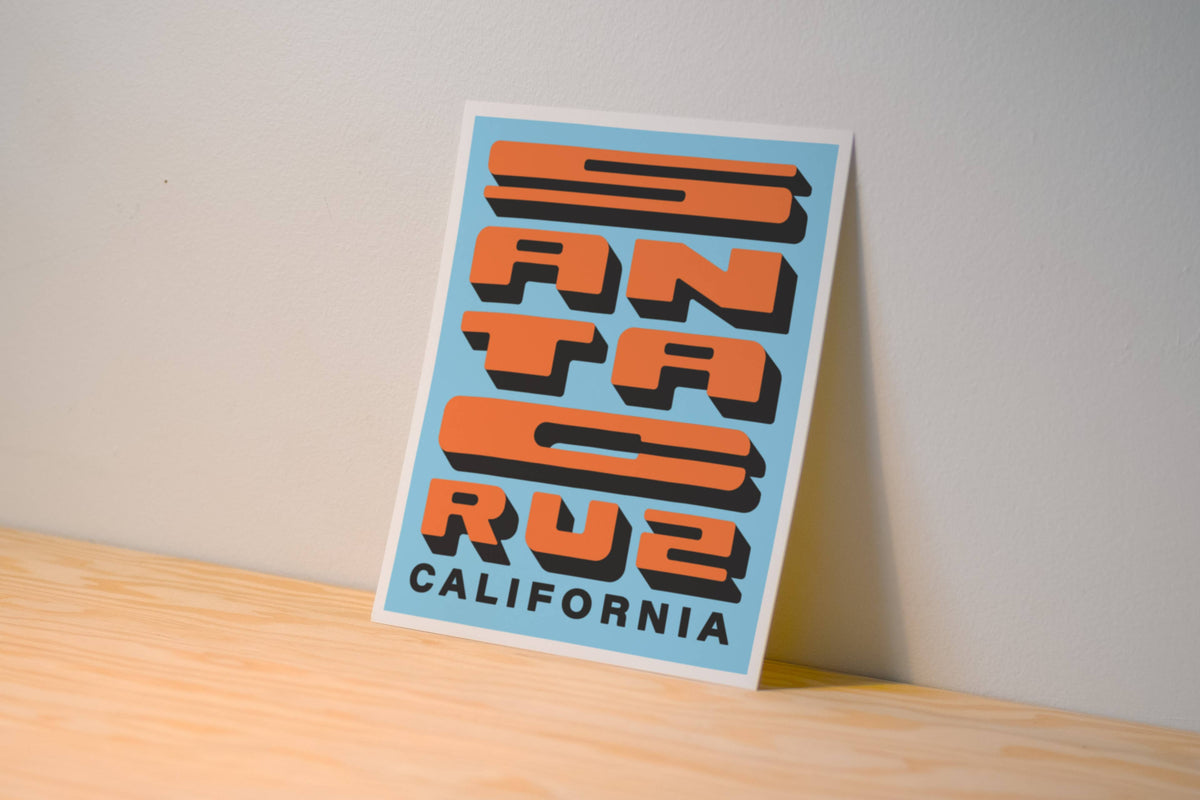A vertical art print featuring a retro-style graphic of the word 'Santa Cruz' in orange and black block letters on a light blue background, with 'CALIFORNIA' printed below. The print is shown leaning against a neutral wall on a light wood surface.