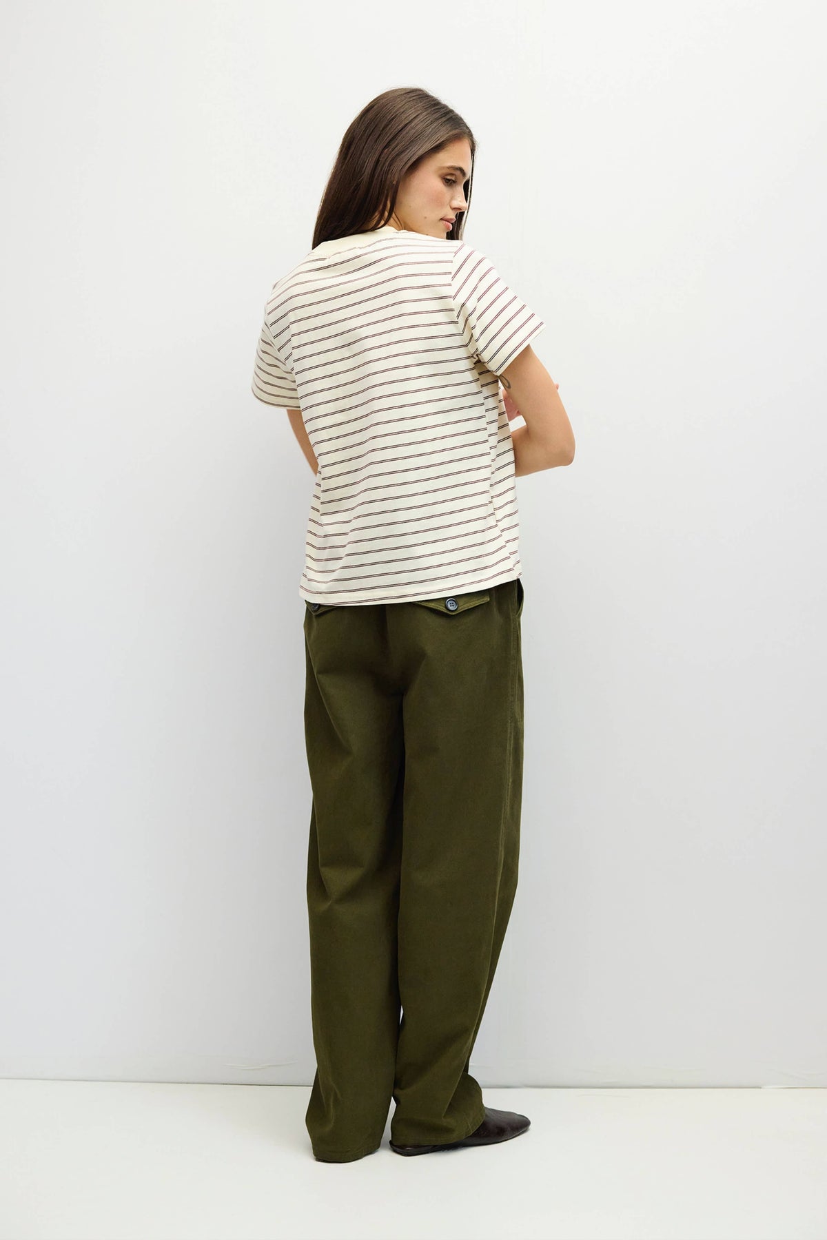 A woman is shown from the back wearing a cream-colored short-sleeve t-shirt with thin dark horizontal stripes and olive green wide-leg trousers, standing against a plain white studio background.