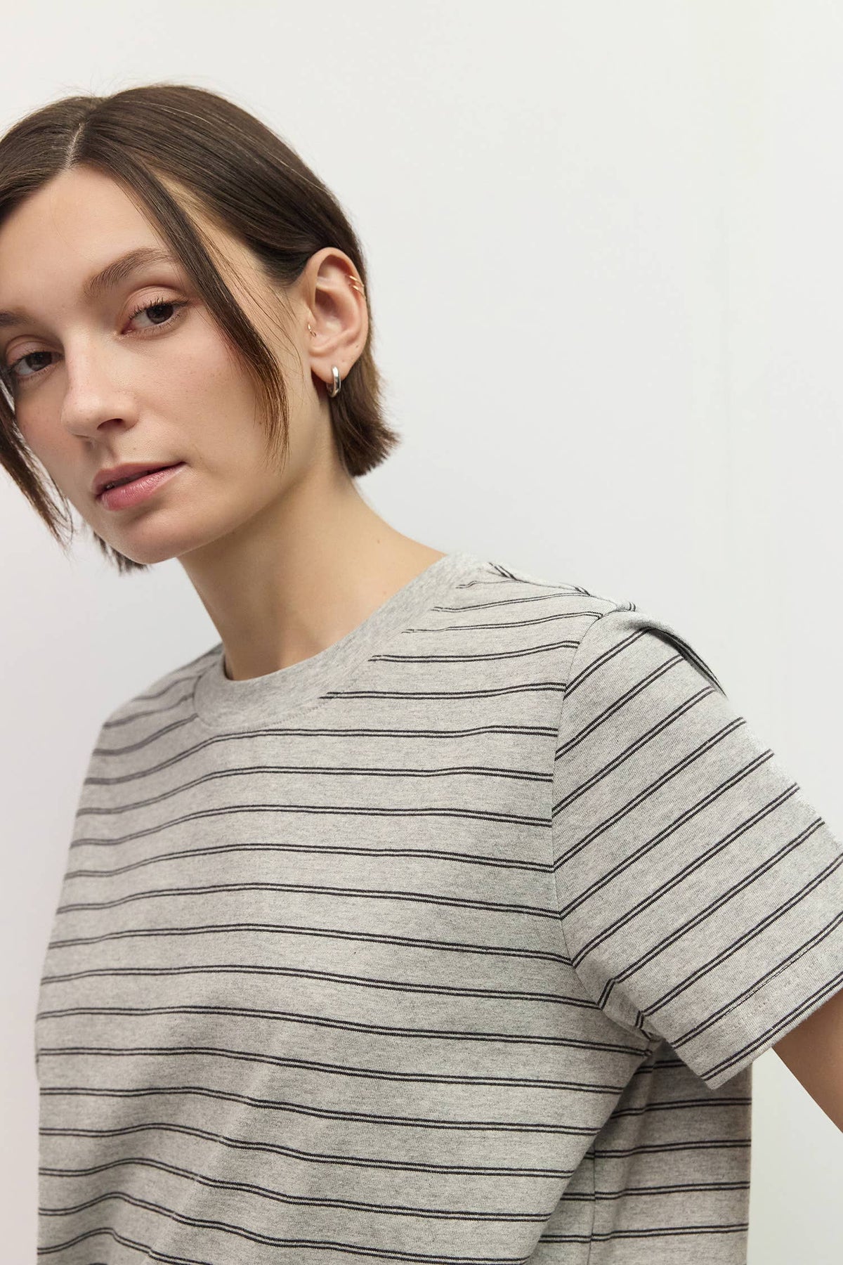 A close-up of a woman wearing a light gray short-sleeve crewneck t-shirt with thin horizontal black stripes, set against a plain light gray background.