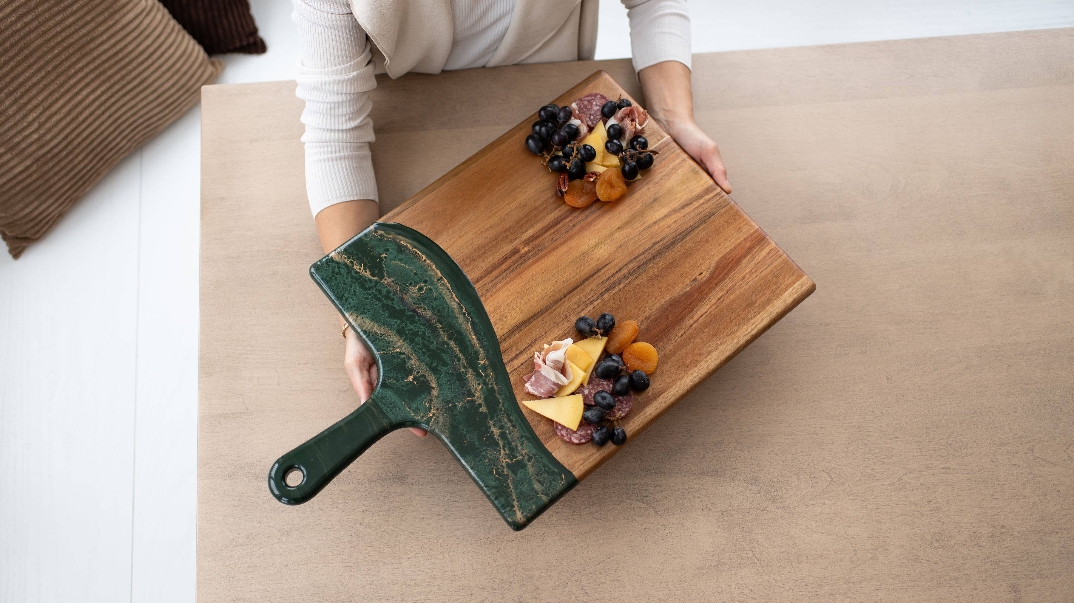 Person holding a wooden cutting board with fruits and a green knife on a light-colored surface.