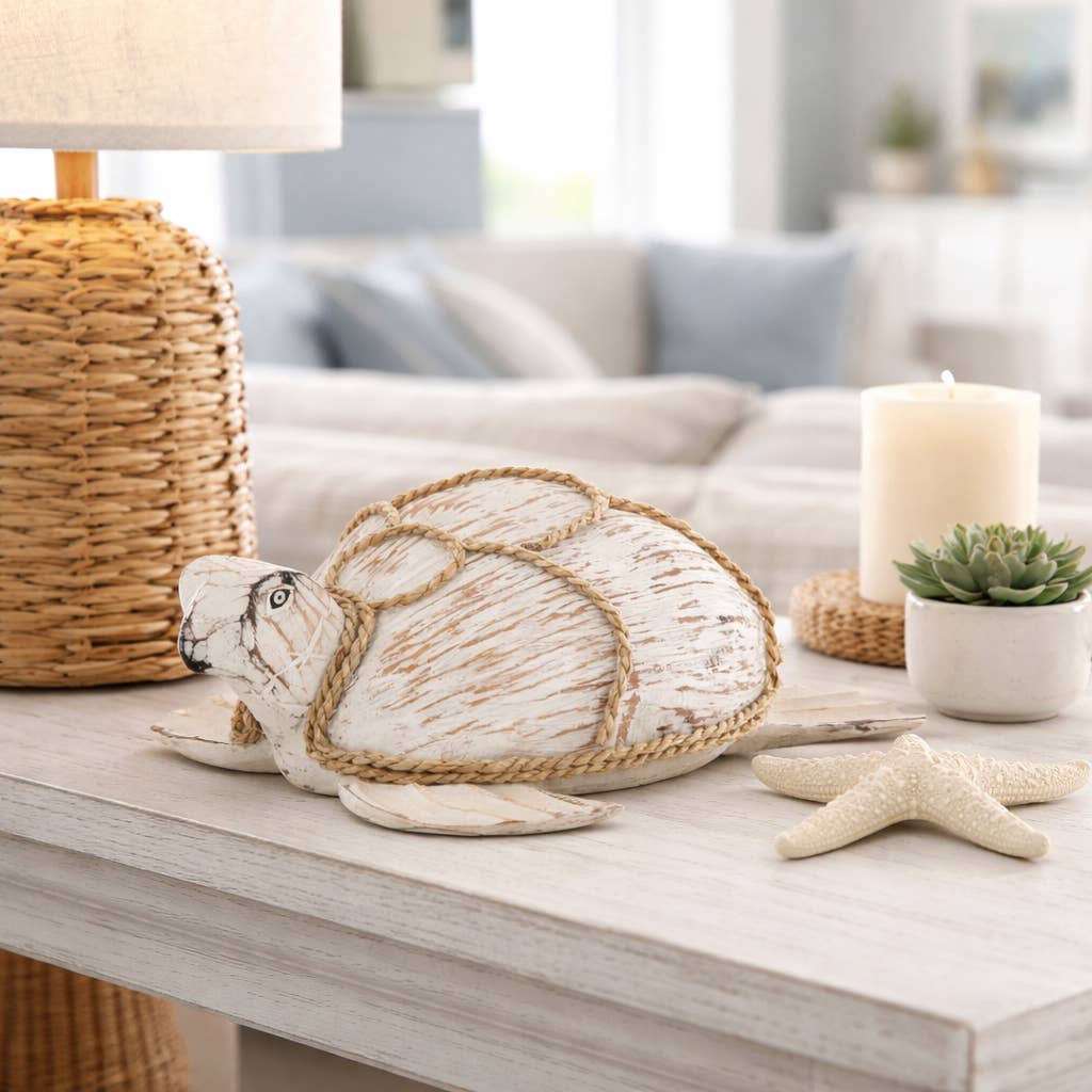 A decorative white and beige sea turtle figurine with rope wrapped around its shell, displayed on a light wood table in a styled living room setting with a lamp, candle, succulent, and starfish.
