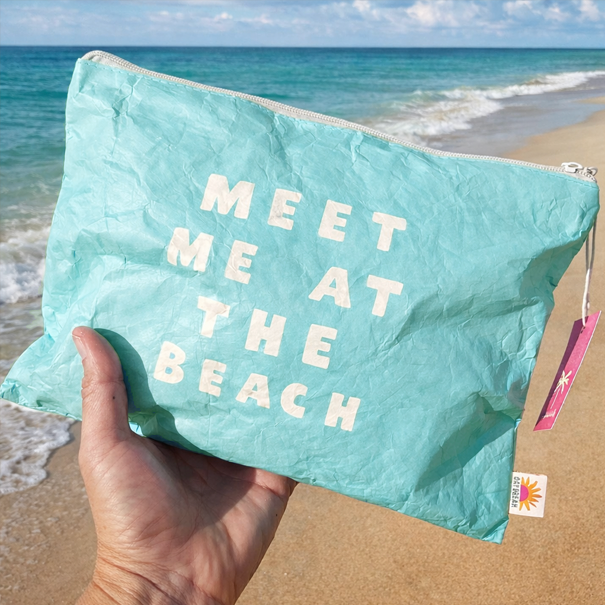 Turquoise pouch with 'Meet Me at the Beach' text held by a hand on a beach.