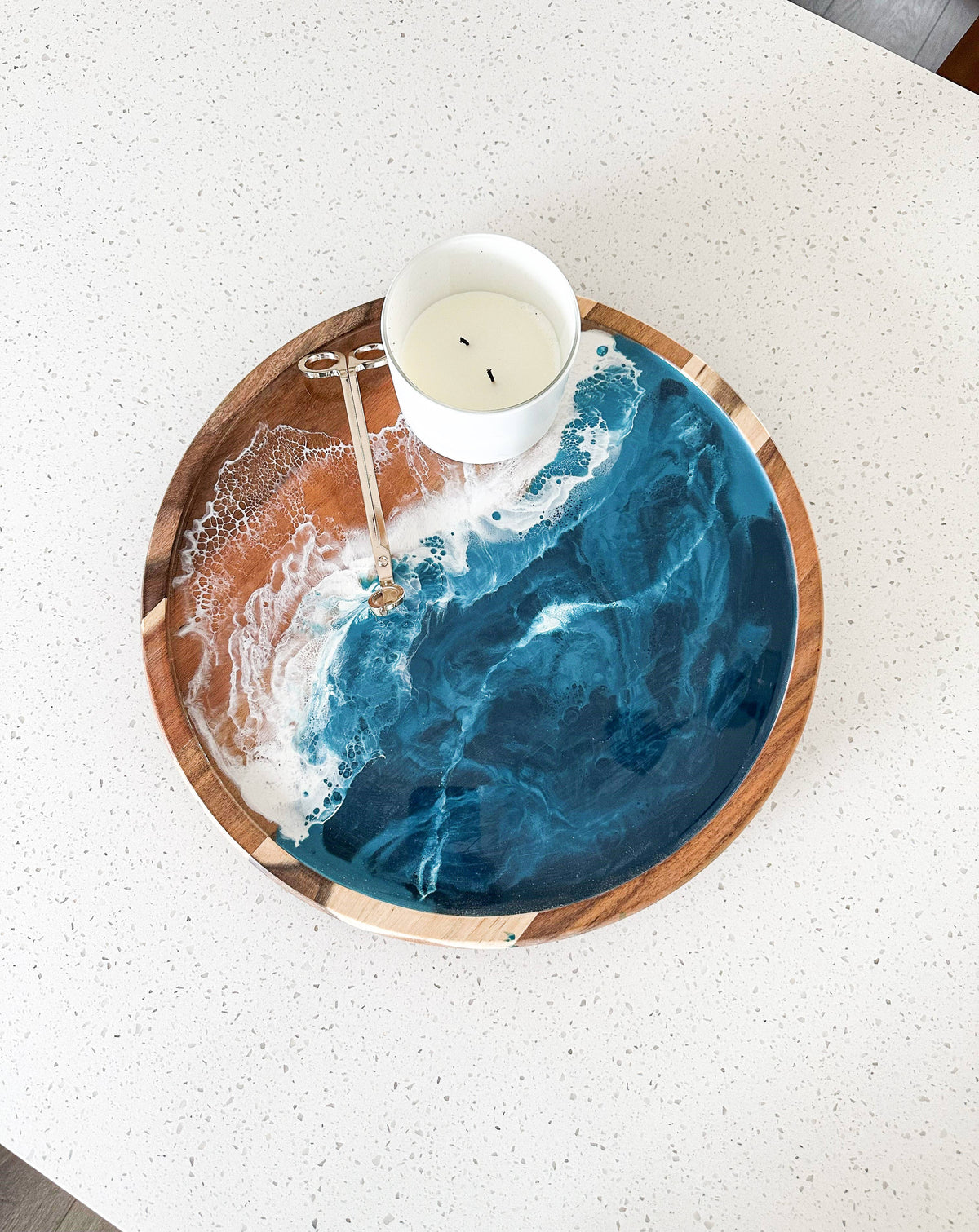 A round wooden serving tray with a blue and white resin ocean wave design, styled on a white speckled countertop with a white candle on top.