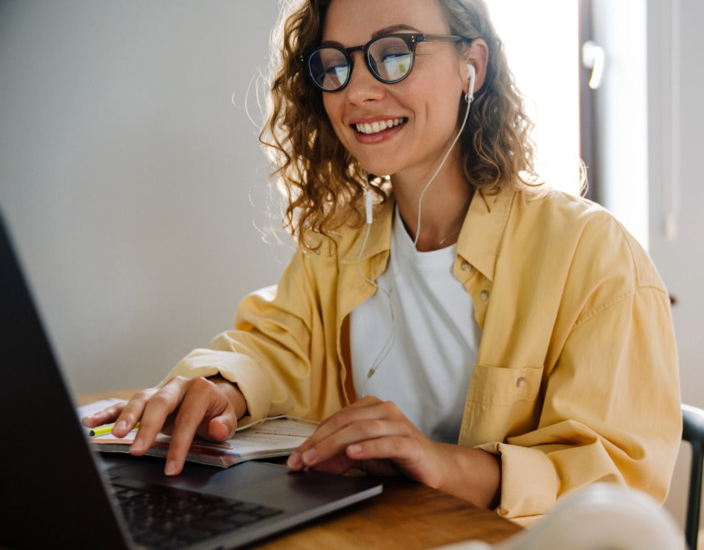 A smiling woman with curly hair and glasses uses a laptop at a desk, wearing a yellow overshirt over a white T-shirt and white earphones.