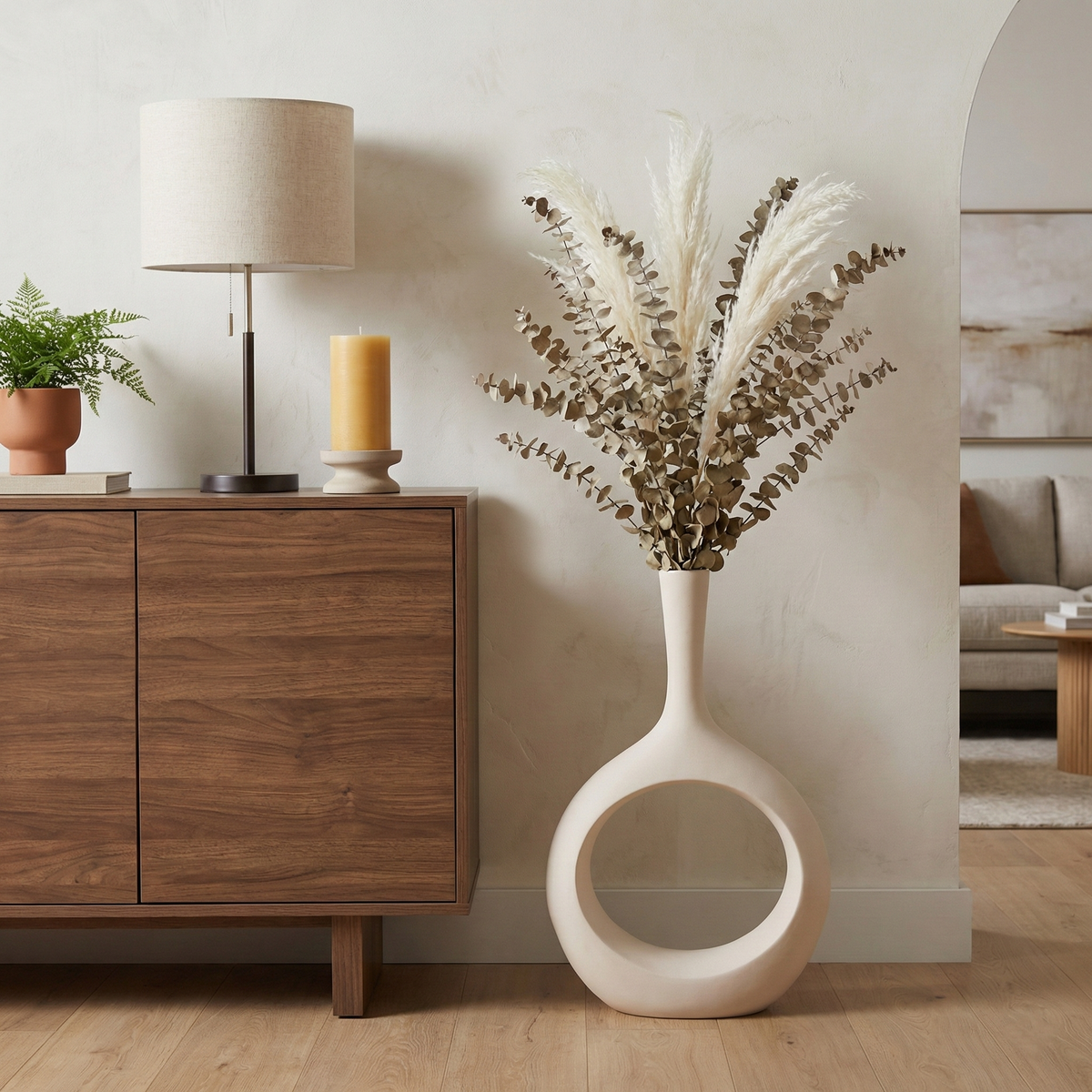 A tall sculptural ceramic vase with a circular cutout, filled with dried pampas grass and eucalyptus, styled in a modern living room next to a wooden sideboard.