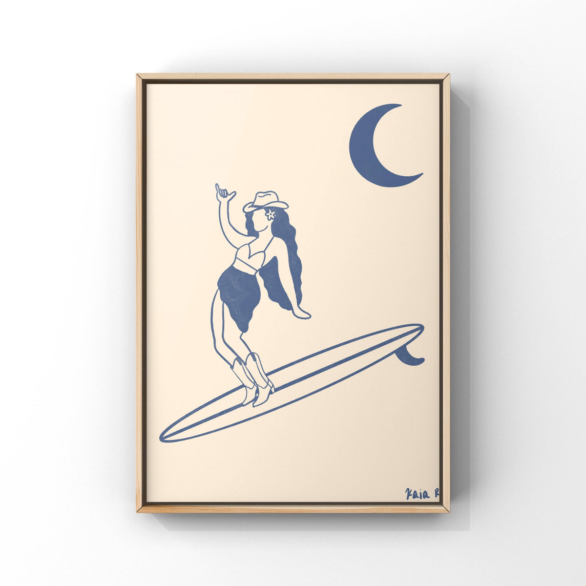 Framed wall art featuring a minimalist navy blue illustration of a woman in a hat and swimsuit standing on a surfboard under a crescent moon, set against a warm cream background with a thin light wood frame.