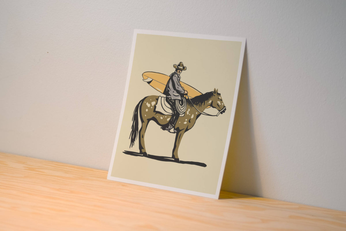A vertical art print featuring a cowboy riding a brown horse while carrying an orange surfboard, displayed leaning against a neutral wall on a light wood surface.