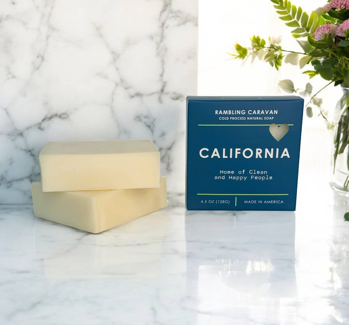 Two pale cream bars of Rambling Caravan California One Hand Wash Natural Soap are displayed on a white marble surface next to their dark blue packaging box, with a floral arrangement in the background.