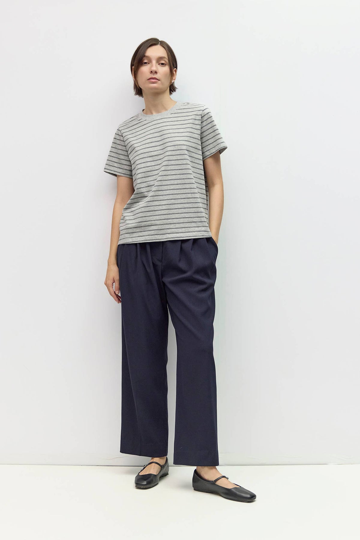A model stands against a plain white studio wall wearing a gray and white horizontally striped short-sleeve t-shirt, dark navy wide-leg trousers, and black flat shoes.