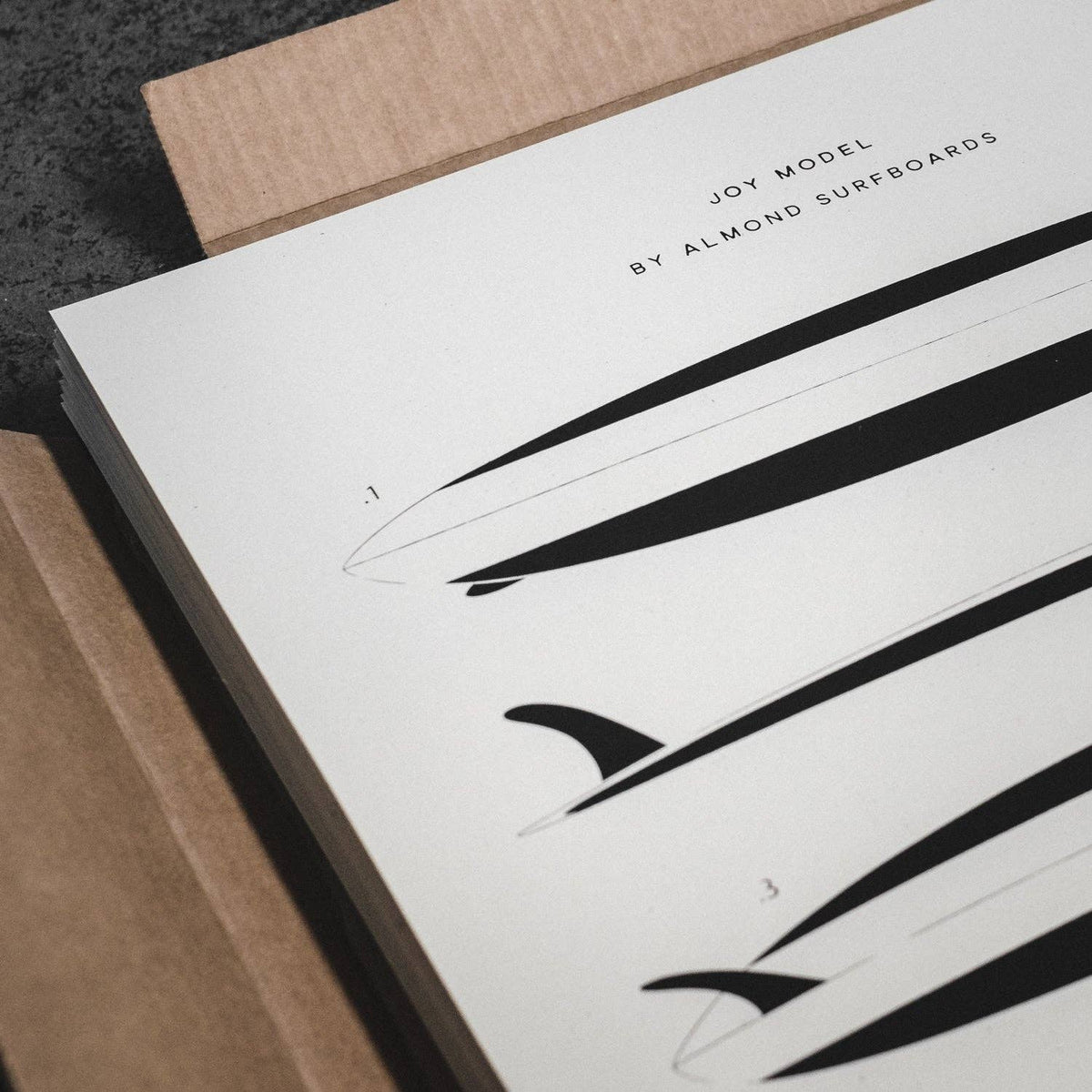 A close-up of a printed surfboard design sheet titled 'JOY MODEL BY ALMOND SURFOARDS' showing black surfboard silhouette graphics on white paper, partially layered over brown kraft paper boxes.