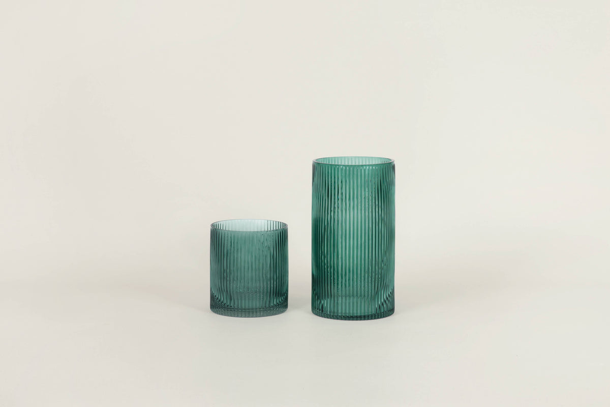 Two ribbed glass tumblers in a translucent teal green color, one small and one larger, standing on a light beige surface against a neutral off-white background.