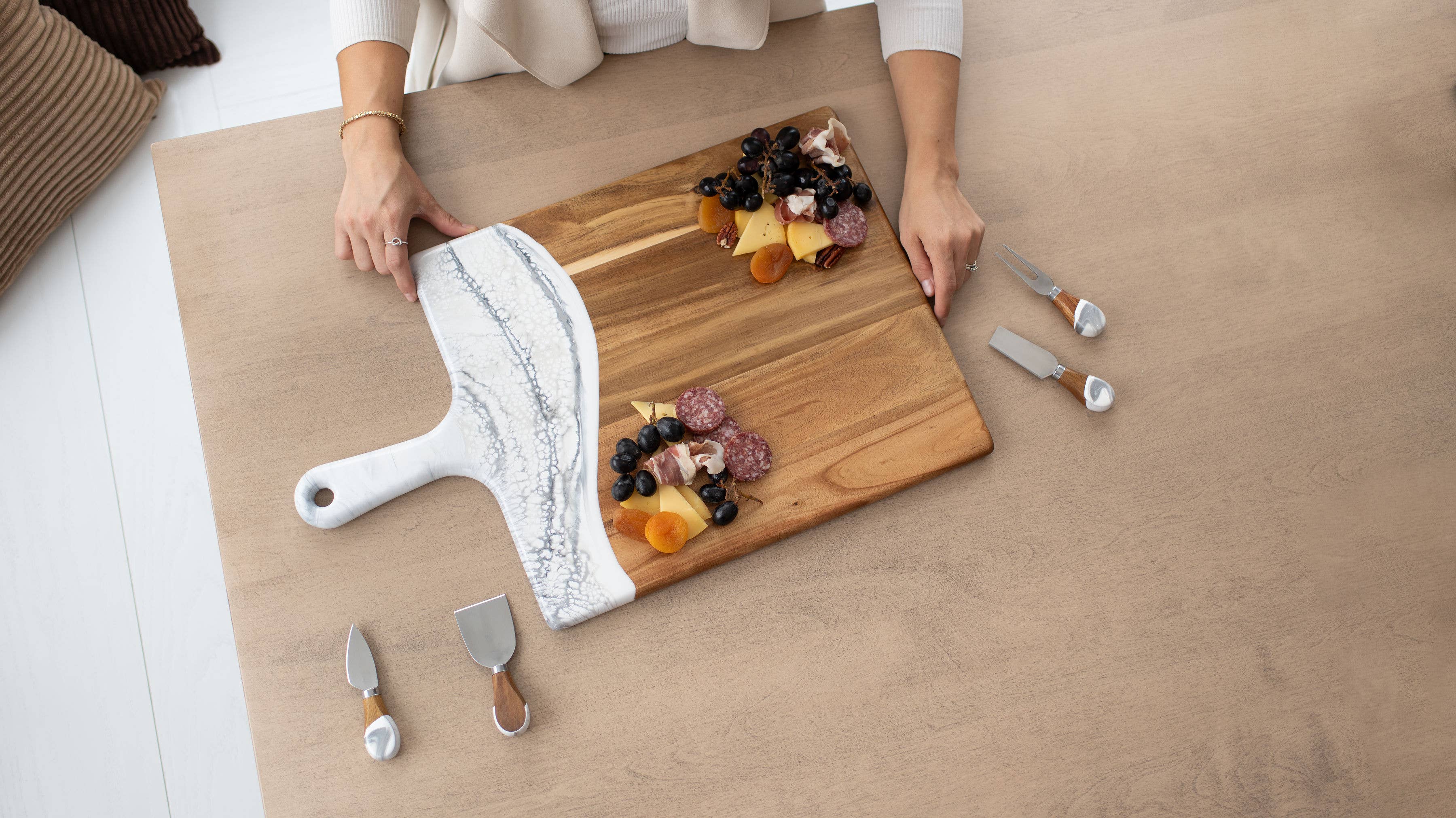 Person arranging dried fruits on a wooden cutting board with cheese knives on a light wood floor.